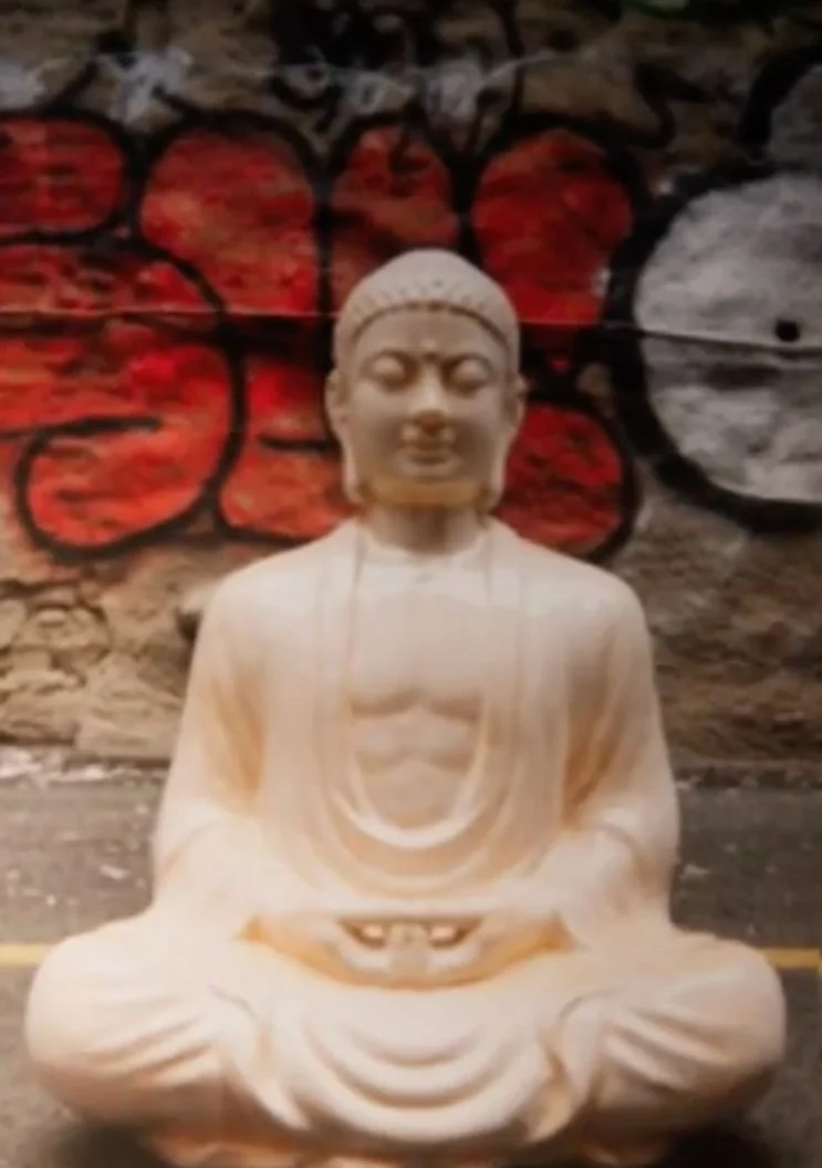 Stone sculpture of a seated meditating figure with a red and black graffiti background.