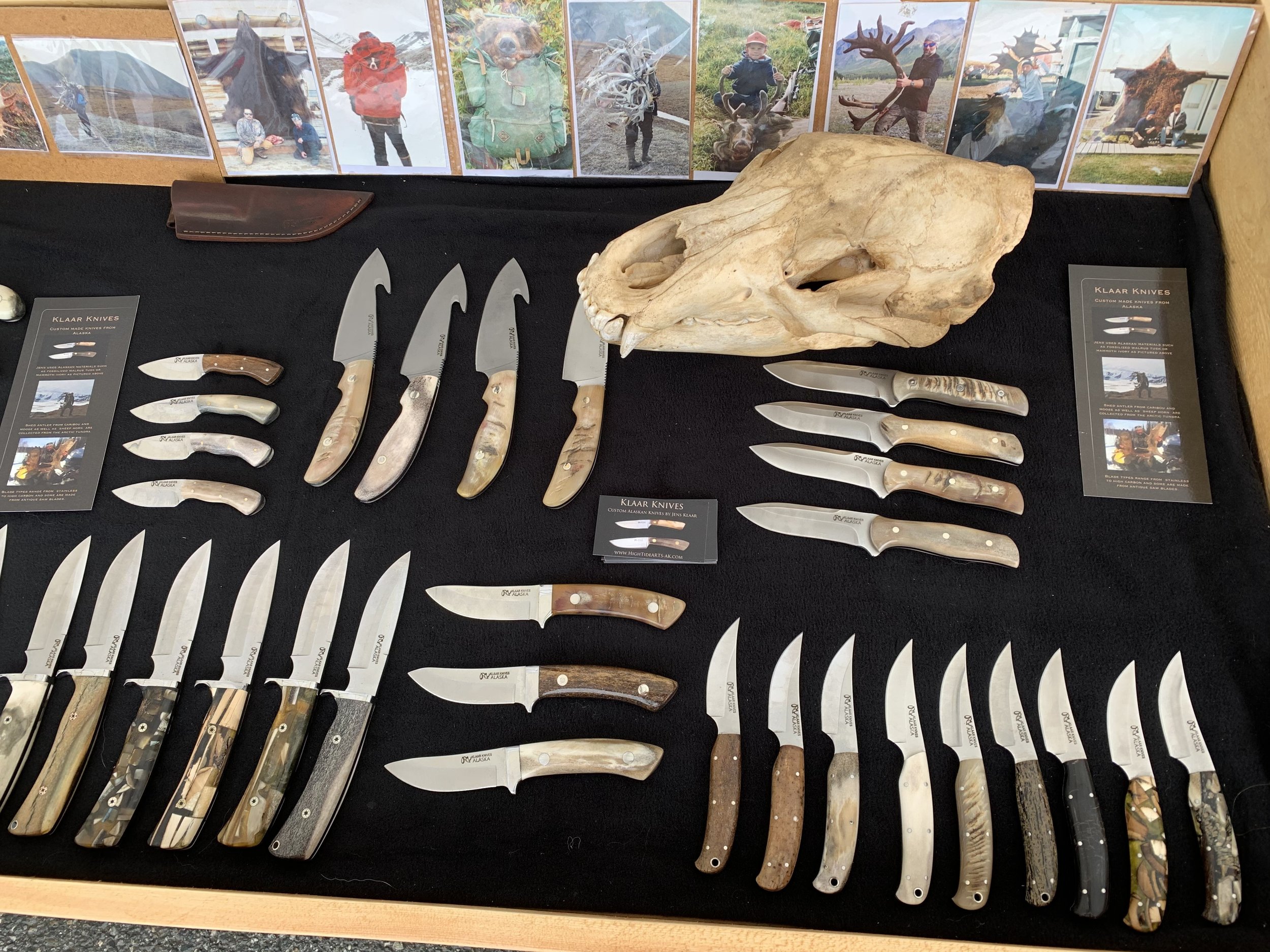 Display of various knives and a large animal skull, with a background of photographs showing people with animals and outdoor scenes. The knives have different handle materials, including wood and mother of pearl, and are arranged on a black surface.