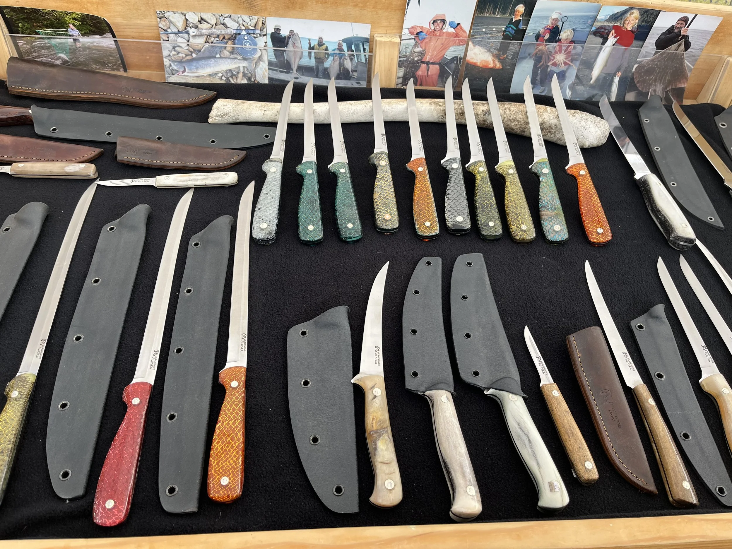 Collection of hunting knives with various colored handles, some with sheath covers, displayed on a black fabric surface with photographs in the background.