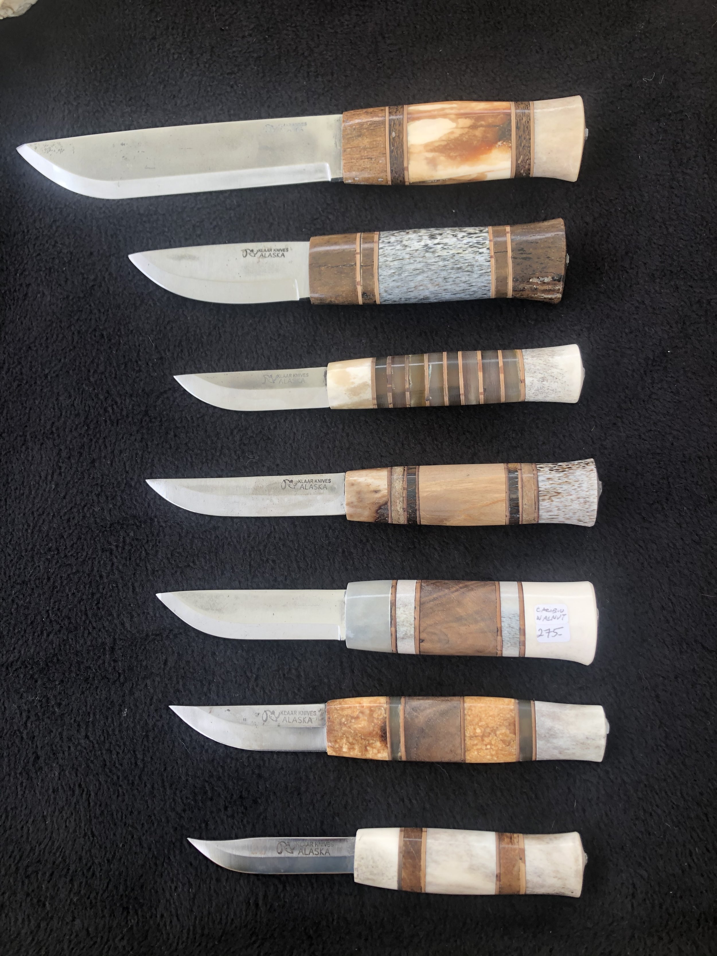Six knives with carved handles, some labeled as Alaska or with price tags, arranged on a black surface.