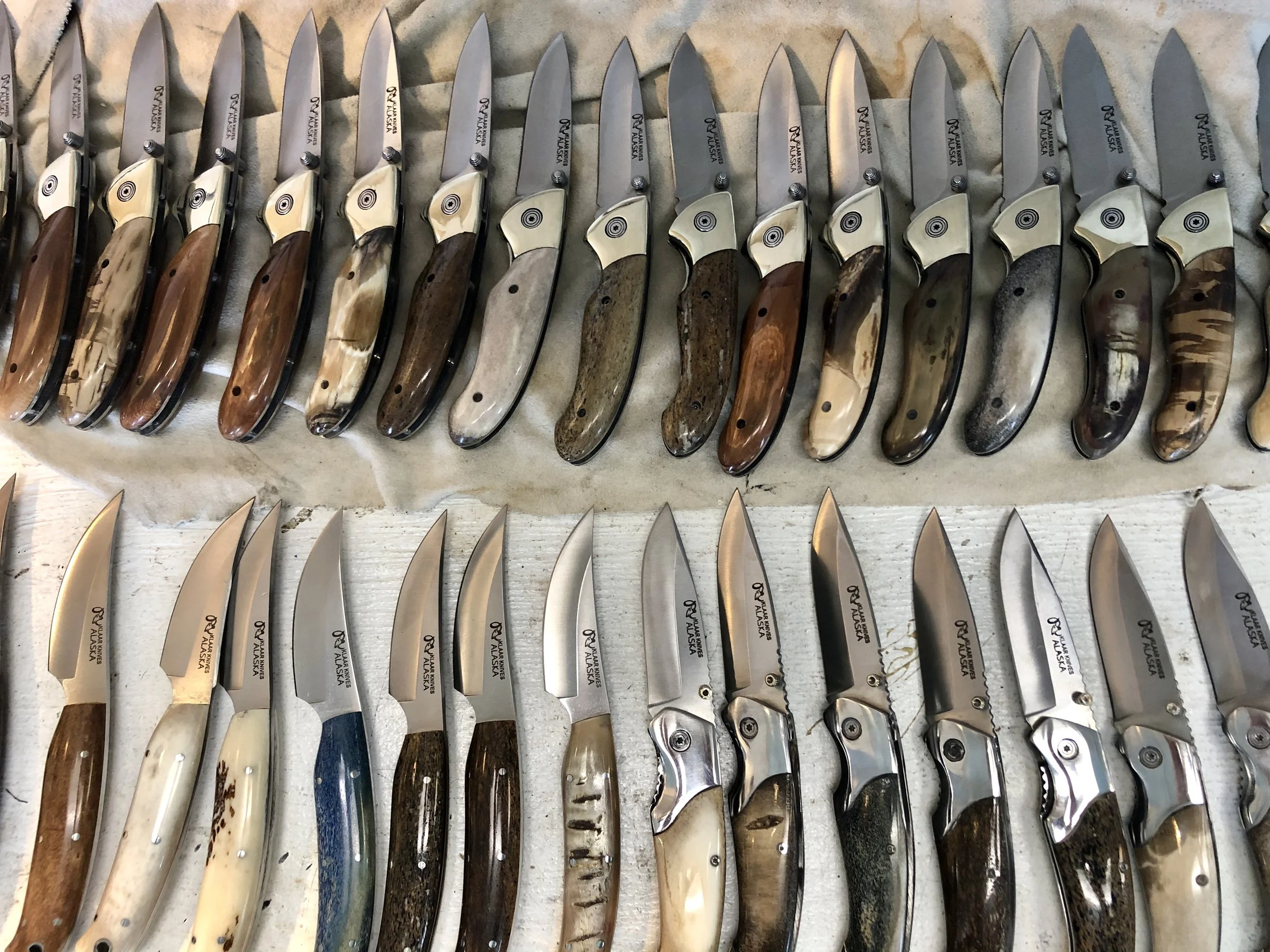 A collection of various folding knives with different handles and blades, arranged in two rows on a fabric surface.