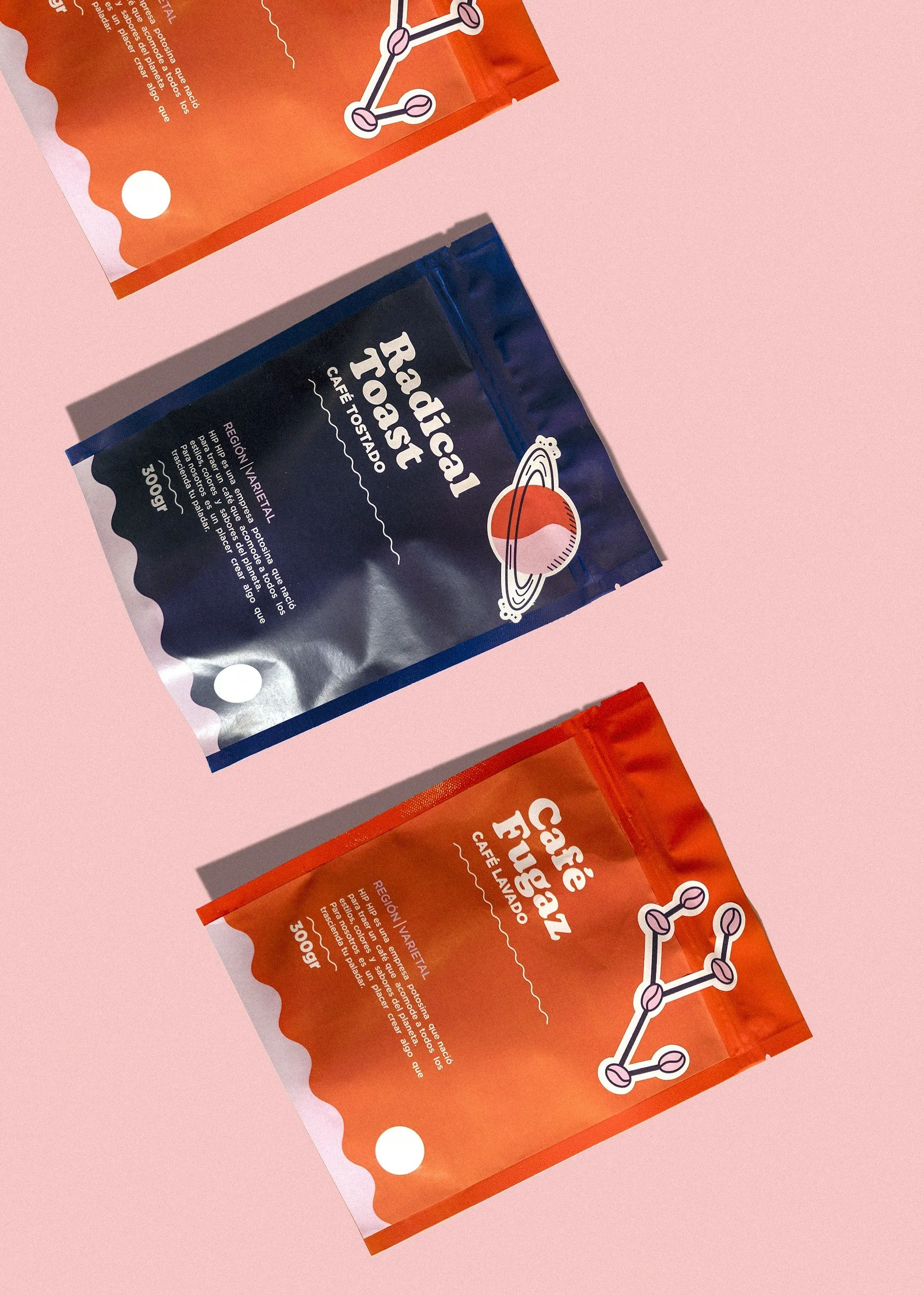 Photograph of two packages of Café Tostado, one in orange and one in black, placed on a pink surface. The packages feature a molecular diagram illustration and text in Spanish describing the product.