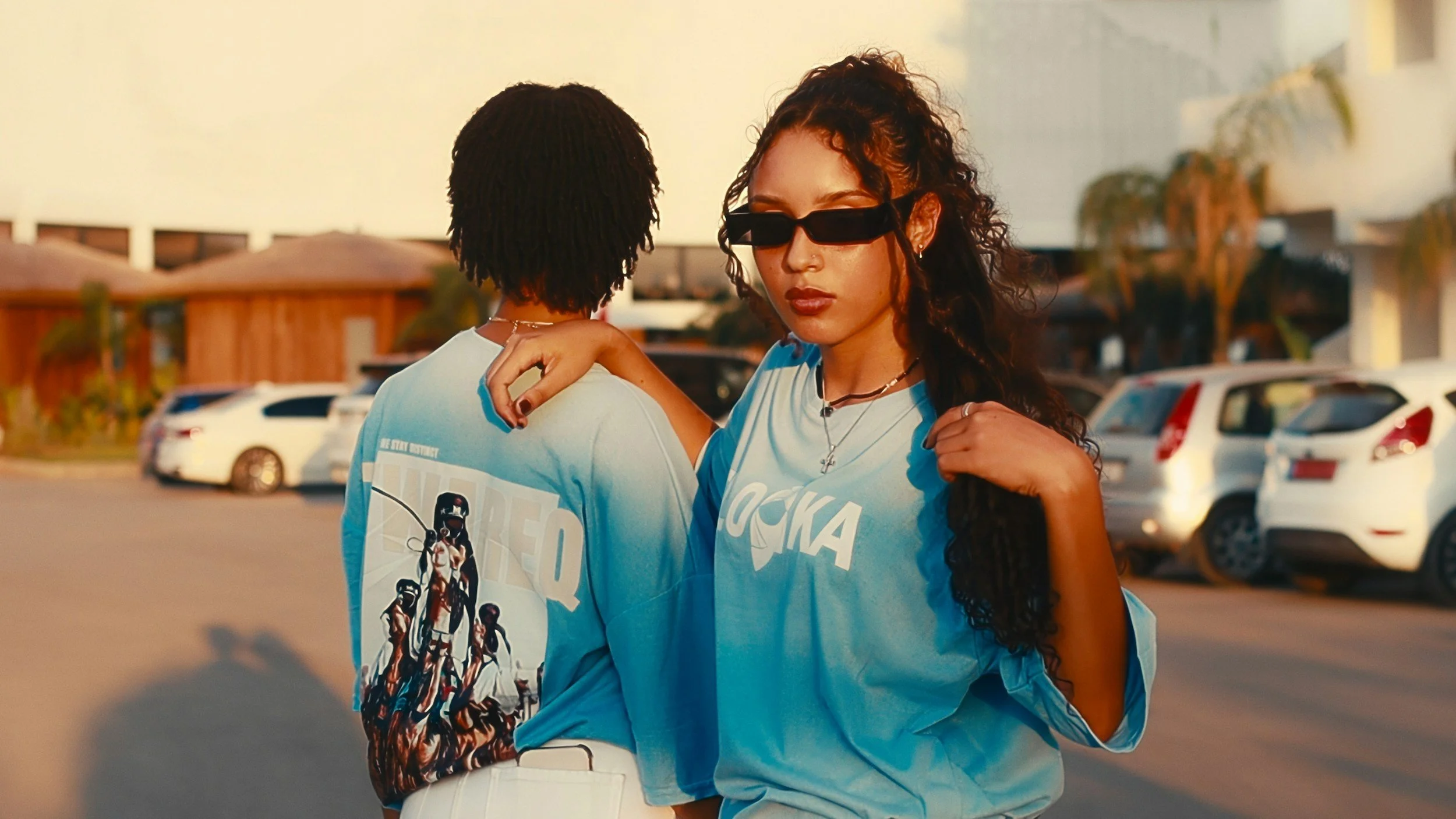 Two young women in matching blue T-shirts standing close together outdoors near parked cars, with one woman's arm around the other's shoulder. One woman has long curly hair and sunglasses, while the other has short hair turned away from the camera.