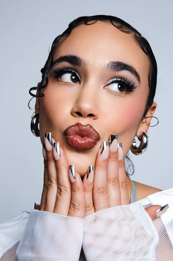 A young woman with dark hair, large hoop earrings, and bold makeup, holding her face with her fingers, pouting her lips, and looking up.