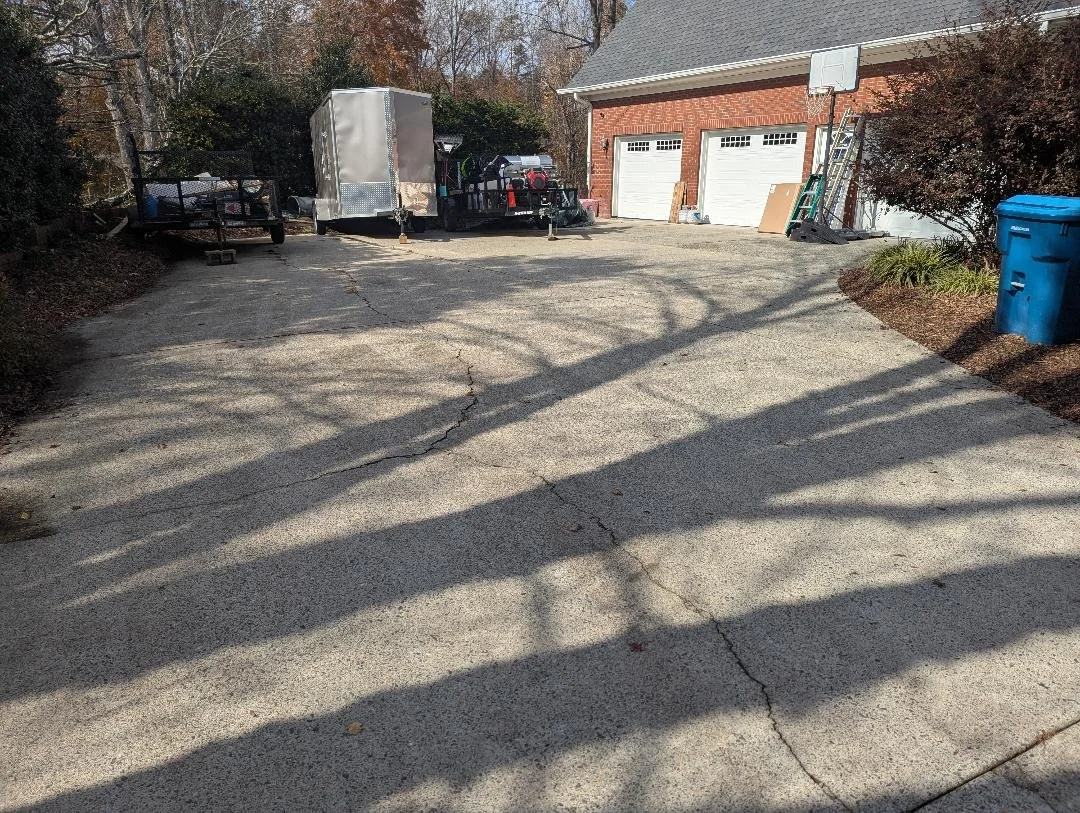 A concrete driveway leading to a brick garage with two white doors. There is a blue trash bin on the right and a ladder leaning against the garage. Several vehicles, including a trailer and a pickup truck, are parked near the garage. Shadows of tree branches are cast on the driveway.