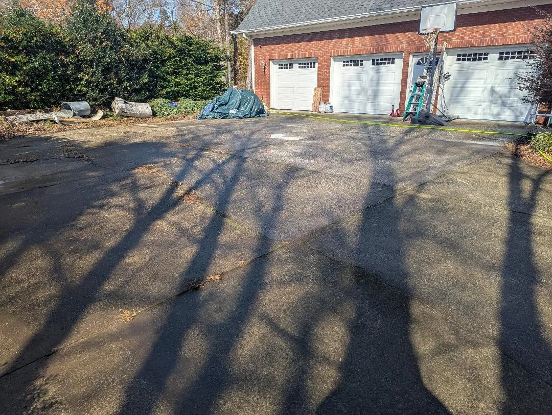 A paved driveway in front of a brick house with a basketball hoop, ladder, and various items near the garage, with leafless trees casting shadows.