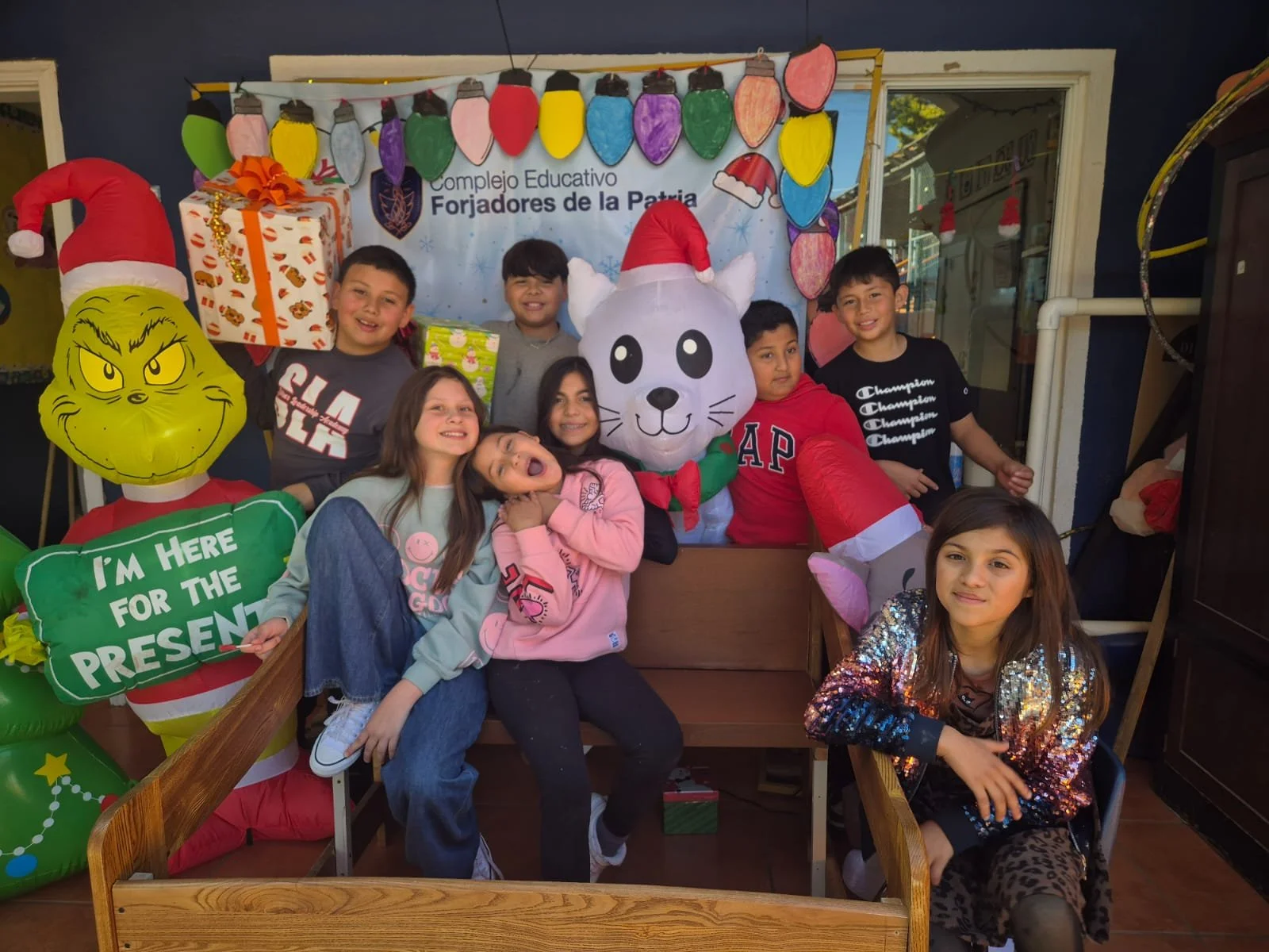 Niños posando con decoraciones navideñas, globos y un grande muñeco de ratón con gorro de Santa en un entorno escolar.