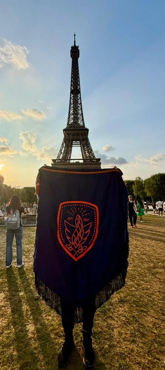 Persona con un escudo azul oscuro y un diseño de llama rojo, de espaldas en primer plano frente a la Torre Eiffel en París, Francia, durante el atardecer.
