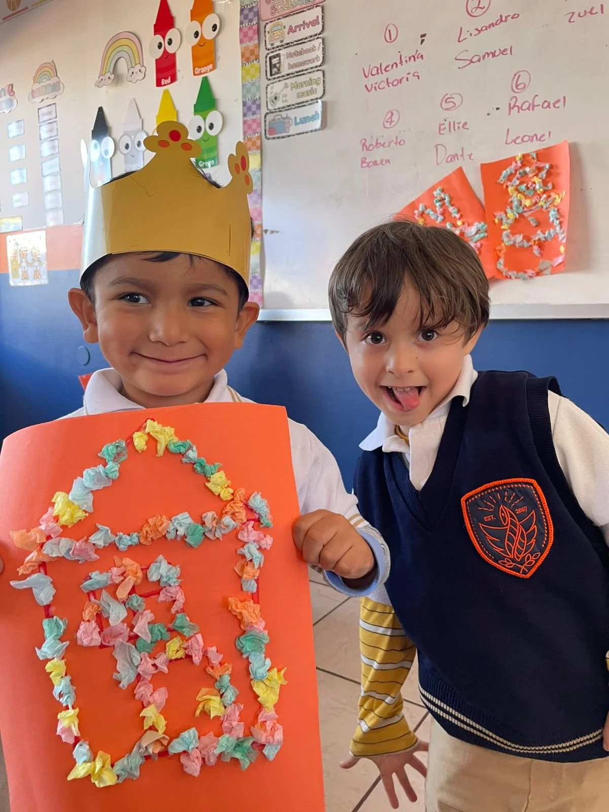 Dos niños en un aula, uno lleva una corona de papel y sostiene una cartulina con un número 8 decorado con pequeños trozos de papel de colores, ambos parecen felices.