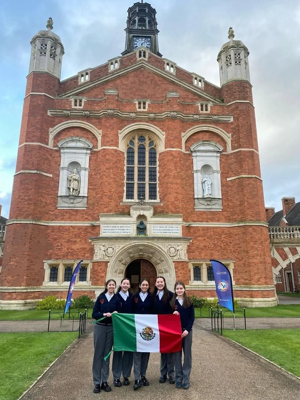 Un grupo de seis jóvenes en uniforme escolar sostiene una bandera de México frente a un edificio de ladrillo rojo con detalles arquitectónicos blancos y una torre en la parte superior. El fondo muestra un cielo nublado y dos banderas azules a los lados.