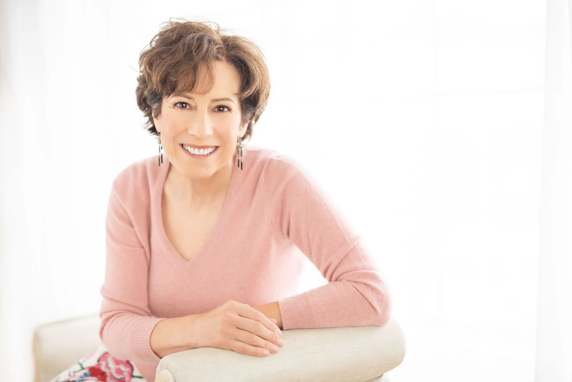 A smiling middle-aged woman with short brown hair, wearing a pink sweater and statement earrings, sitting at a table in a bright, white room.