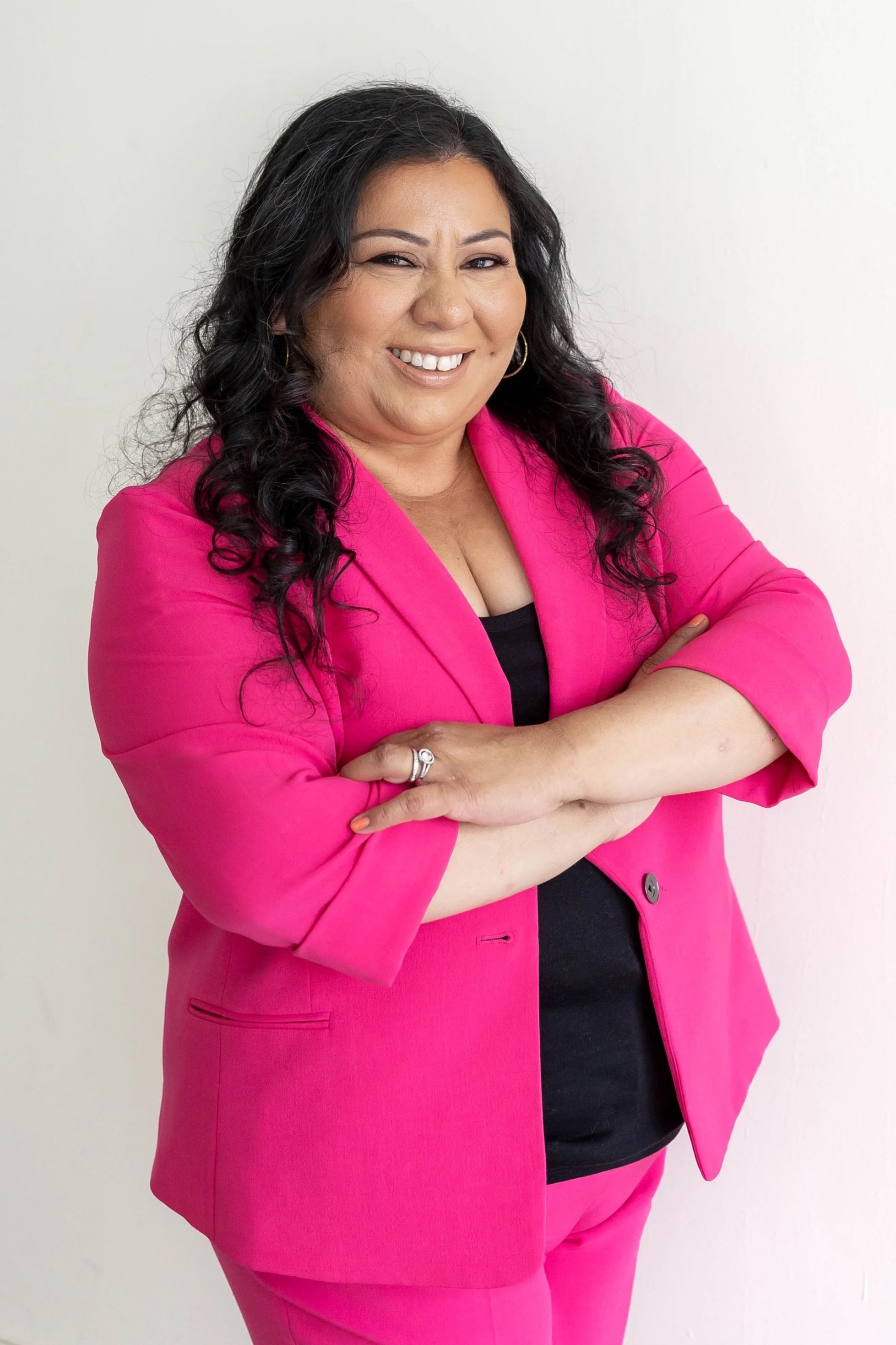 A woman with long black wavy hair smiling and crossing her arms, wearing a bright pink blazer and matching pants over a black top, standing against a plain light background.