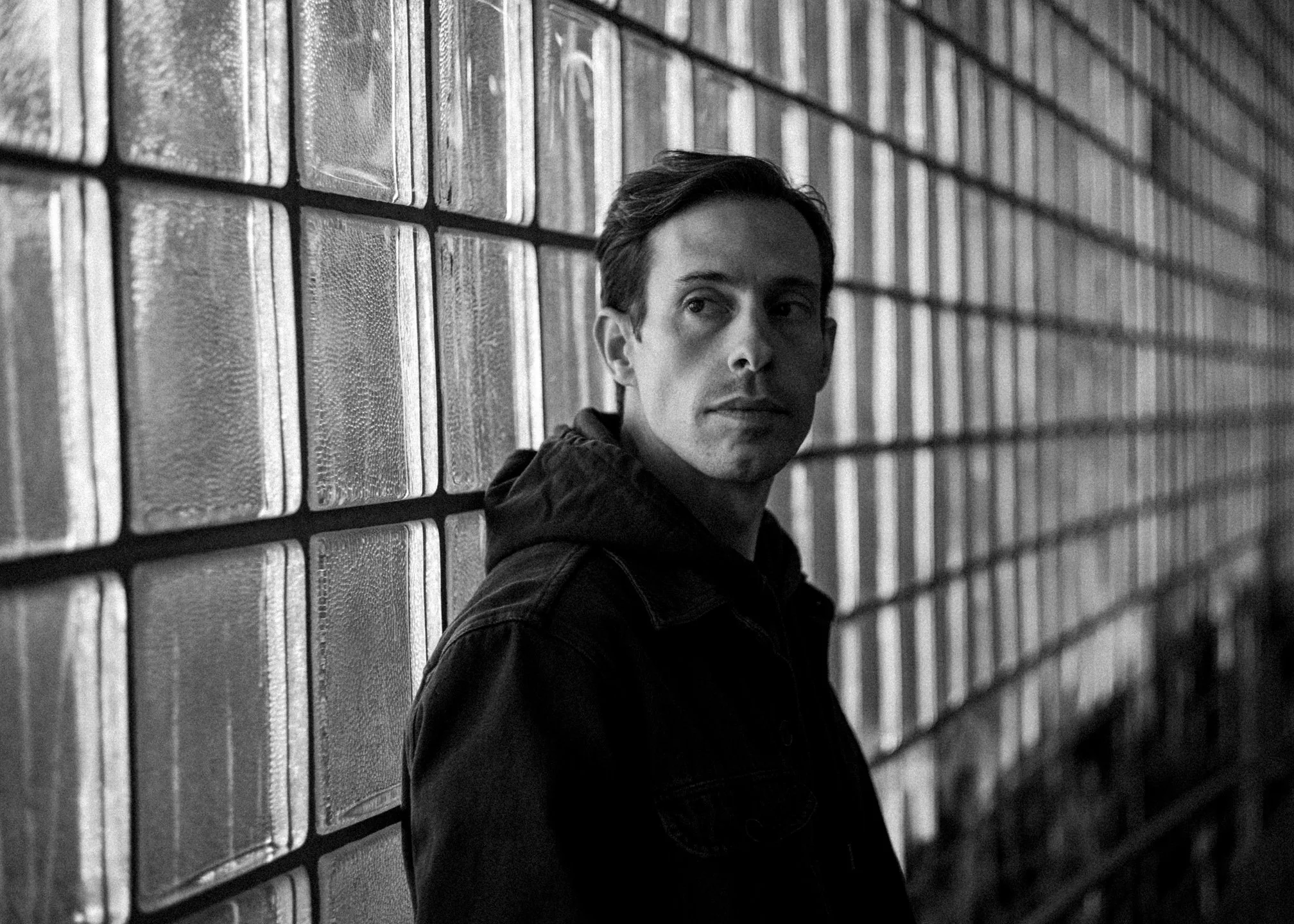 A black and white photo of a man with short hair, wearing a dark jacket, leaning against a textured brick wall with glass blocks, looking to the side.