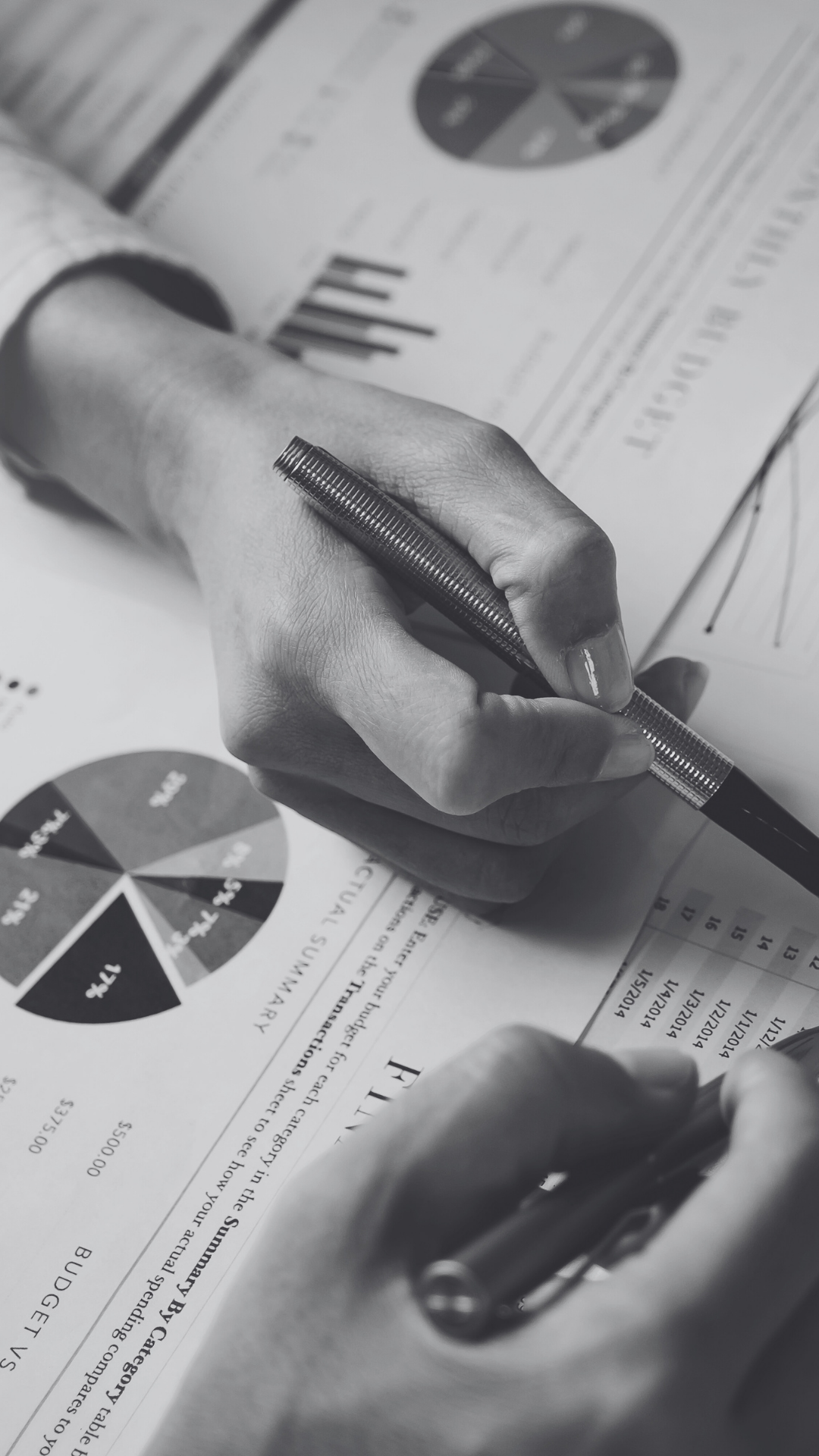 Black and white photo of a person’s hand holding a pen over financial documents with pie charts and bar graphs.
