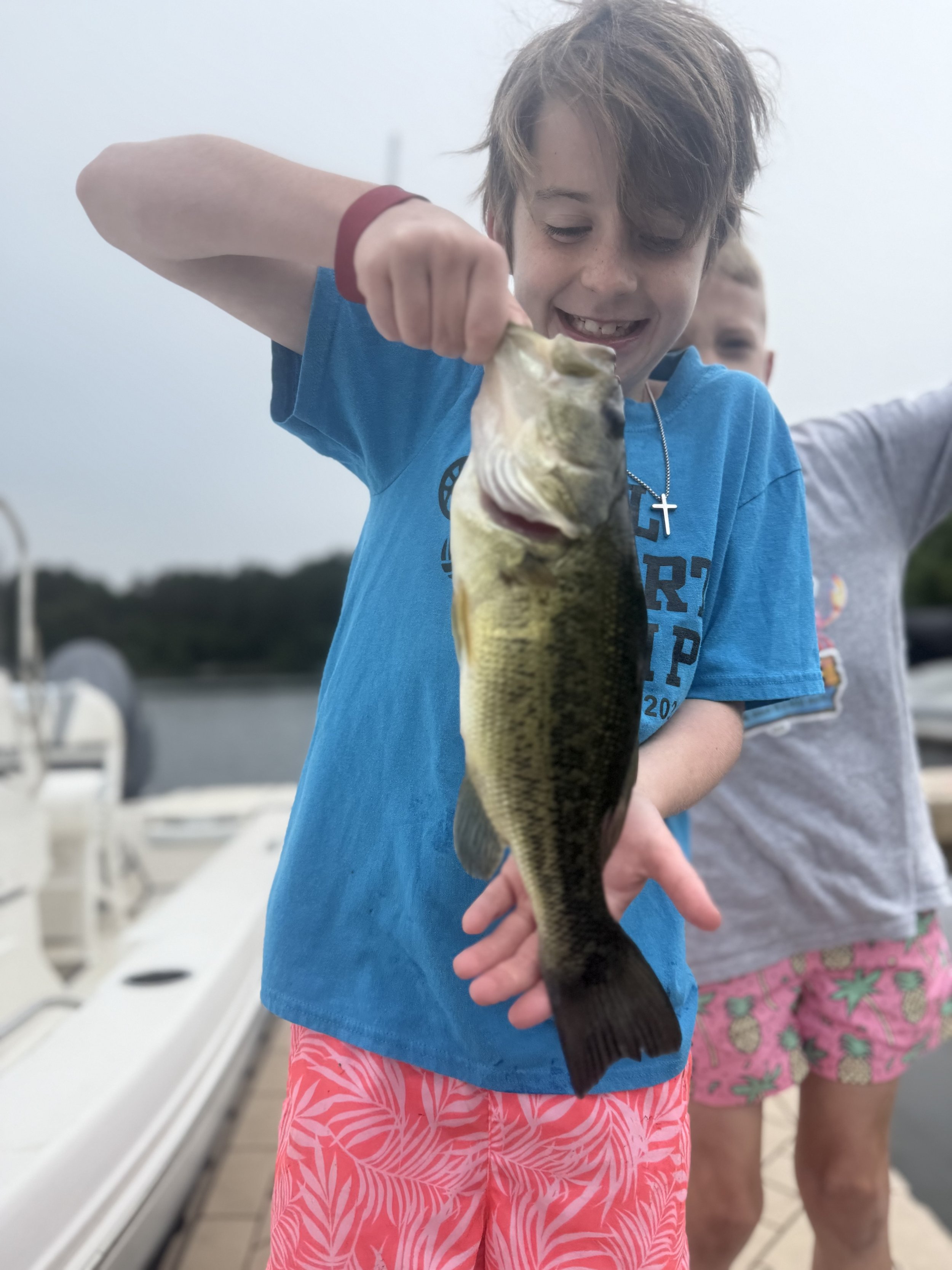 A boy holding a large fish on a boat dock, smiling. There is another child in the background. The children are dressed in casual summer clothes.