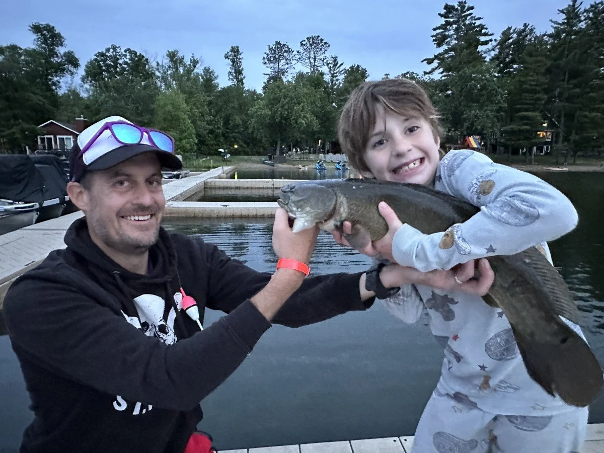 A man and a boy holding a large fish together by a lake during dusk, with trees and some buildings in the background.
