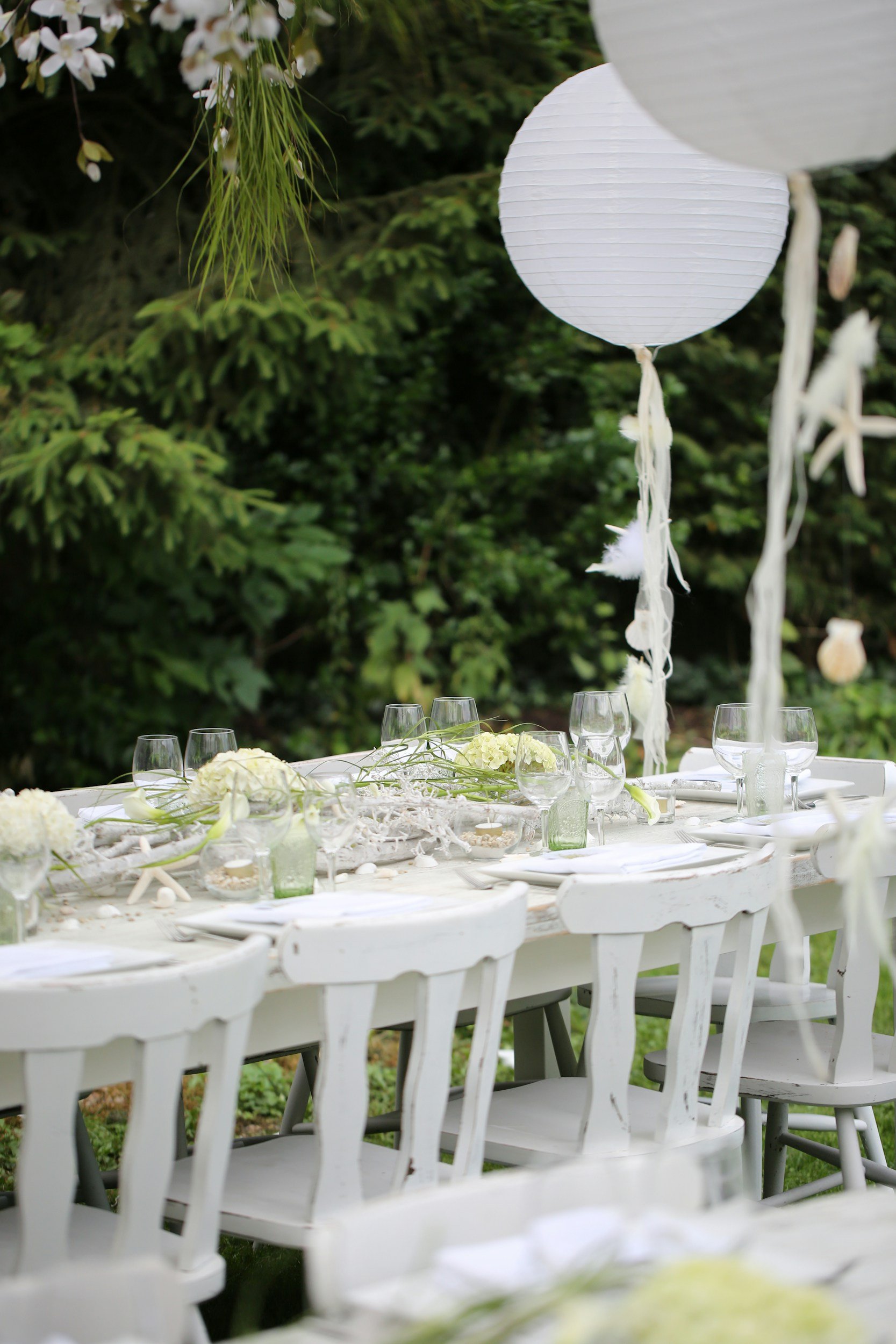 Outdoor table decorated for a celebration, with white chairs, floral centerpieces, glassware, and floating white paper lanterns, set against green foliage.