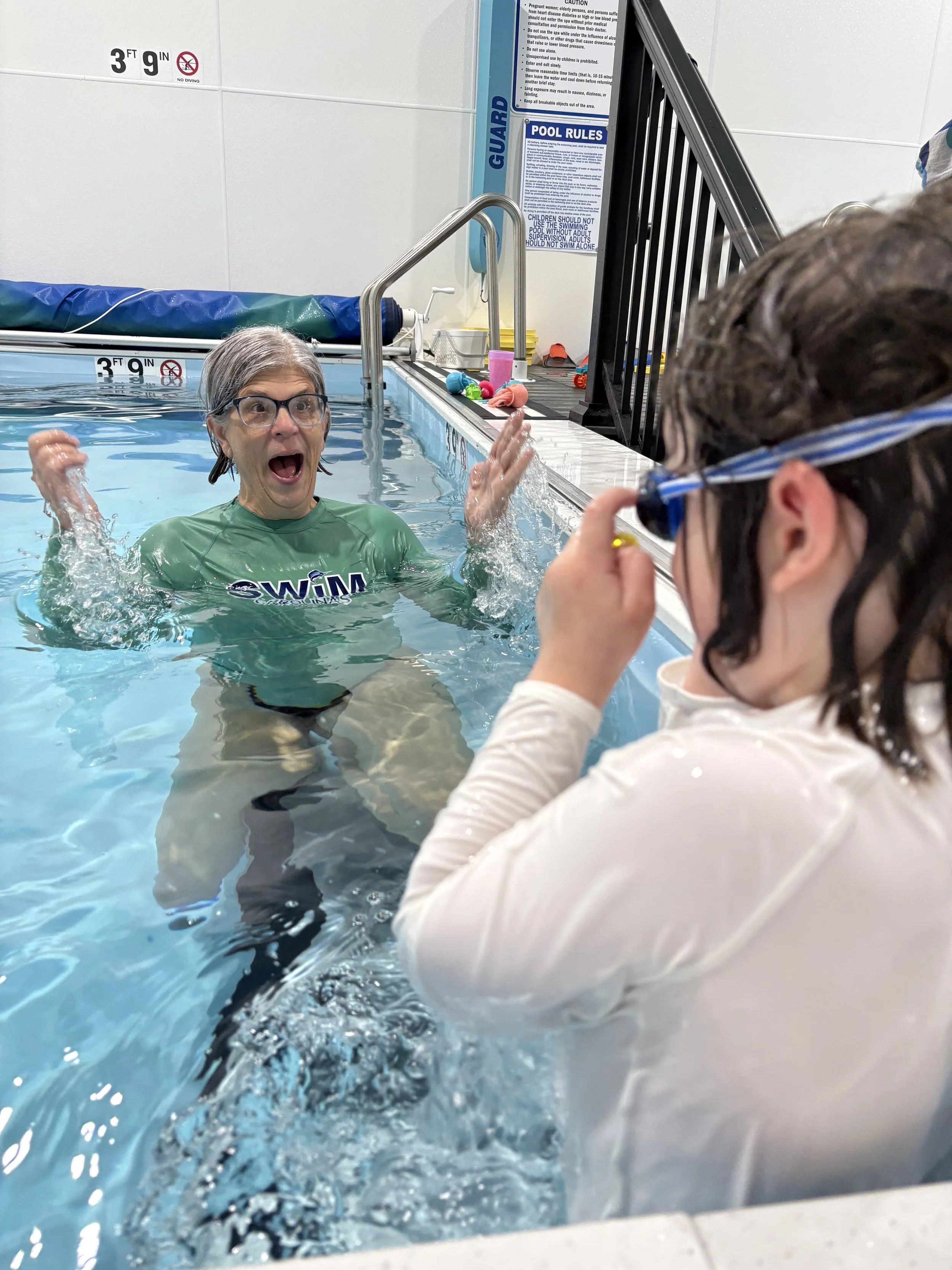 Cathy Sheafor in a green swimming shirt and goggles is excitedly splashing water in a swimming pool, with a child watching her.
