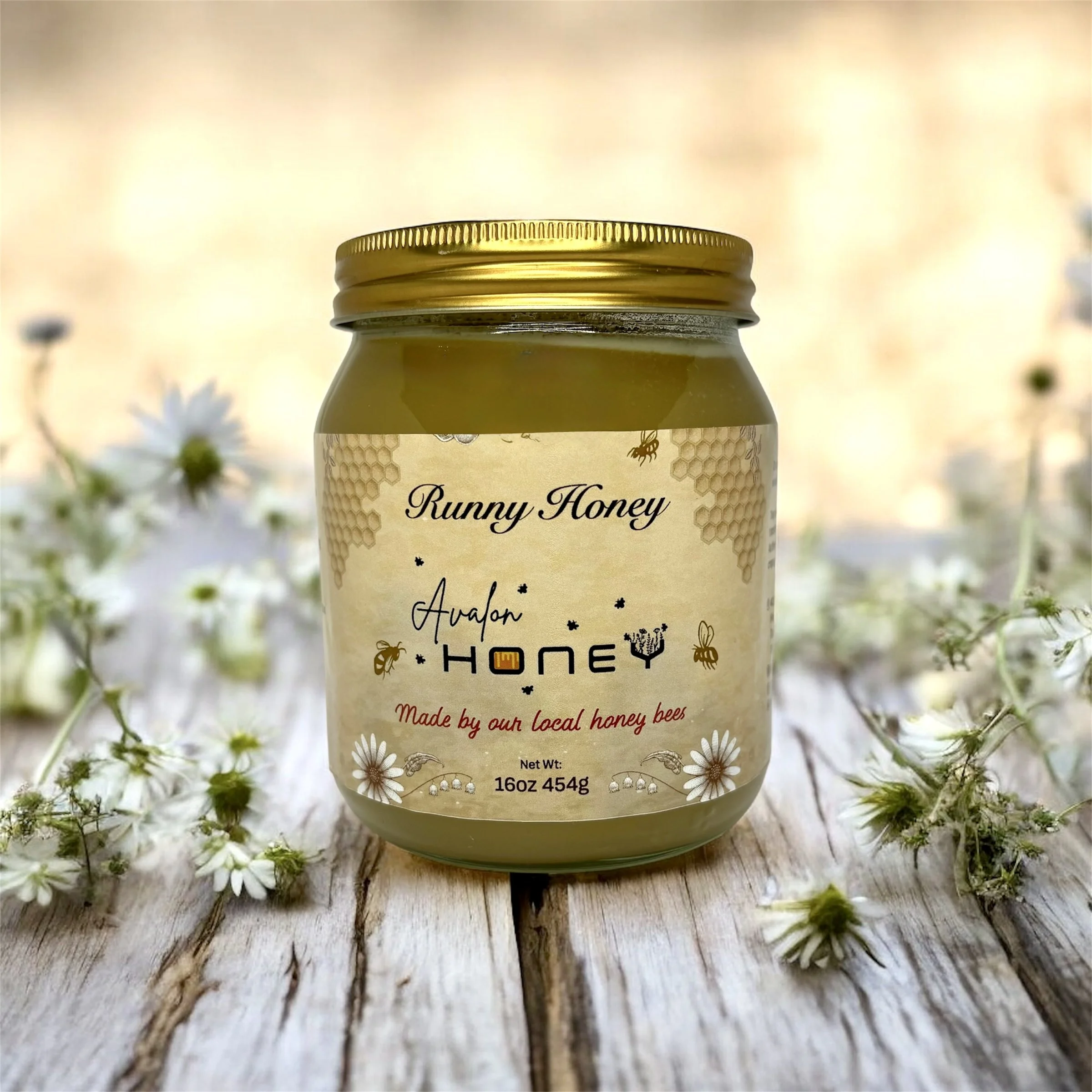 A jar of honey labeled 'Runny Honey Avalon Honey,' with a gold lid, placed on a wooden surface surrounded by small white flowers.