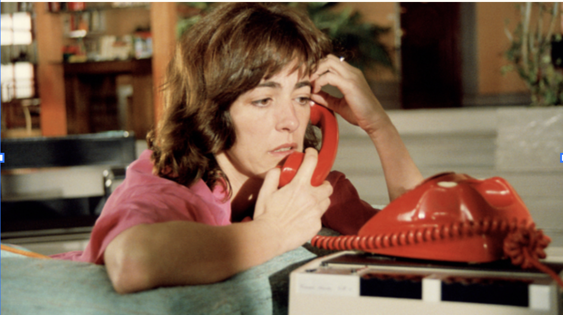 Woman with brown hair and pink shirt lying on her stomach, talking on a red rotary phone in a room with bookshelves.