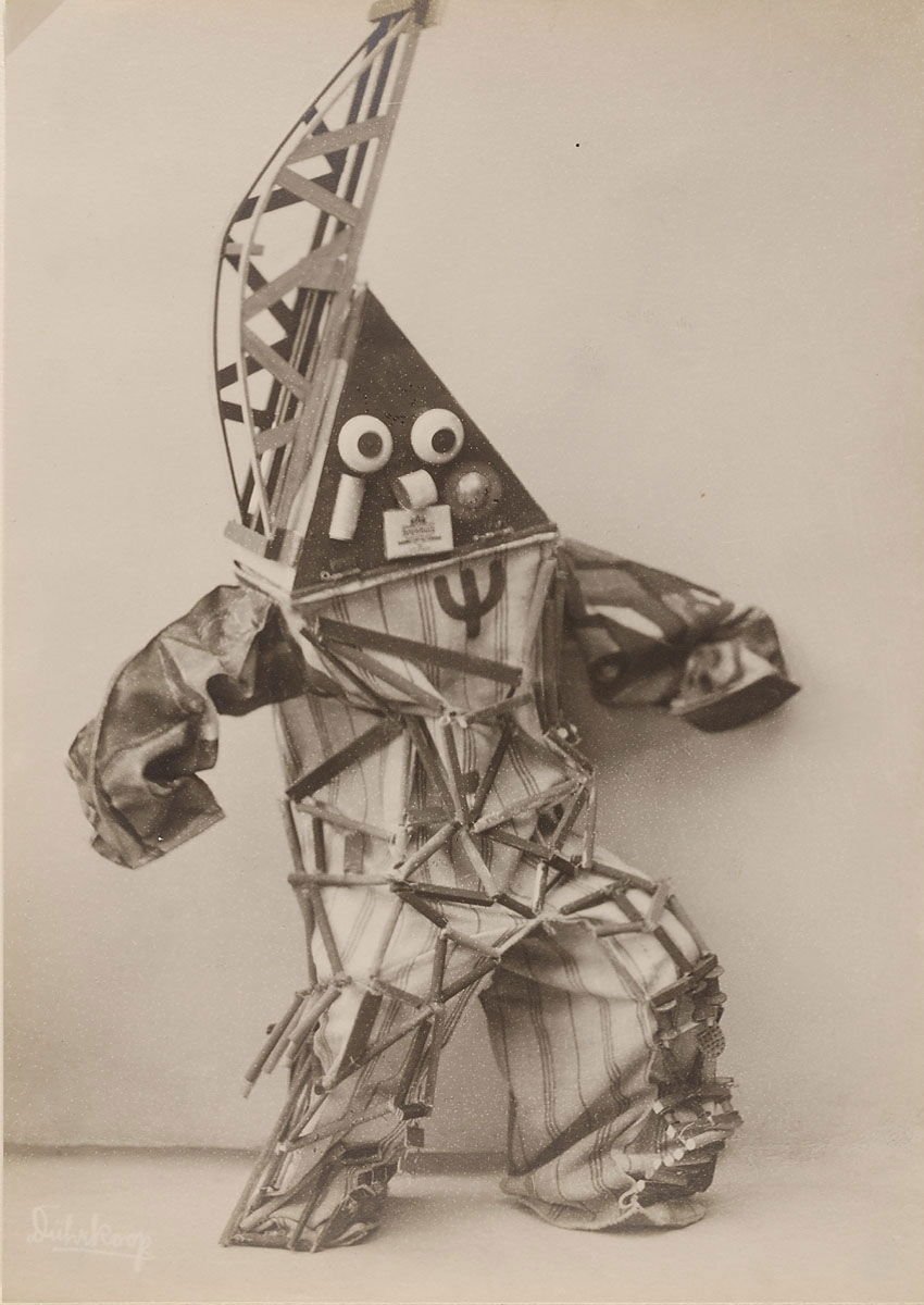 Collage of various objects arranged to resemble a humanoid figure, featuring a head with googly eyes, a torso with a peace symbol, and limbs made from assorted materials, including fabric and metal.