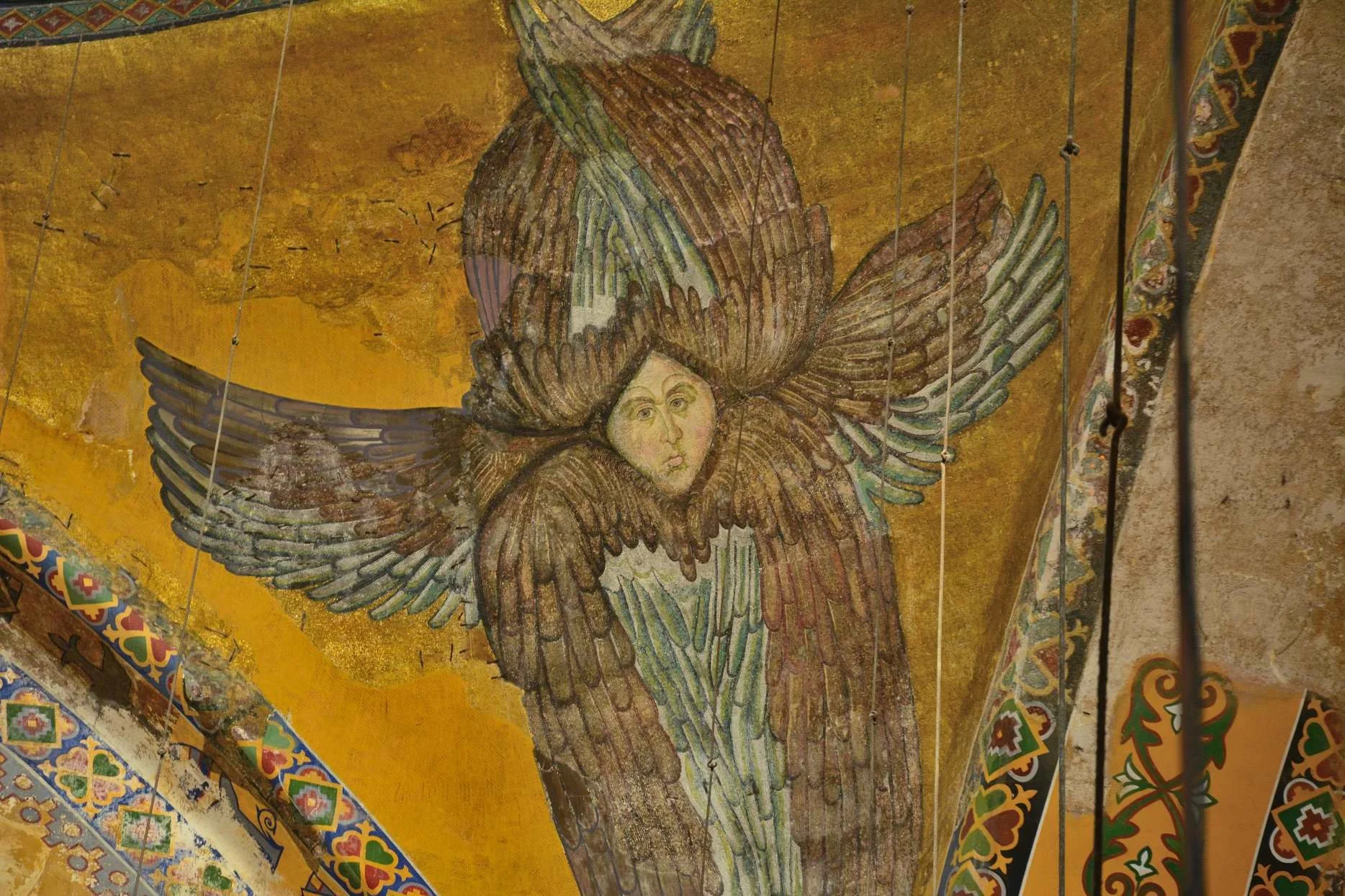 A fresco painting of an angel with large feathered wings and a human face, set against a golden background with decorative patterns along the ceiling edges.
