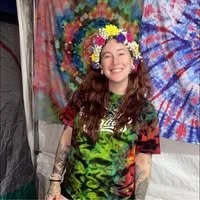 Young woman with long curly hair wearing a flower crown and tie-dye shirt, smiling in front of colorful tie-dye fabric backdrops.