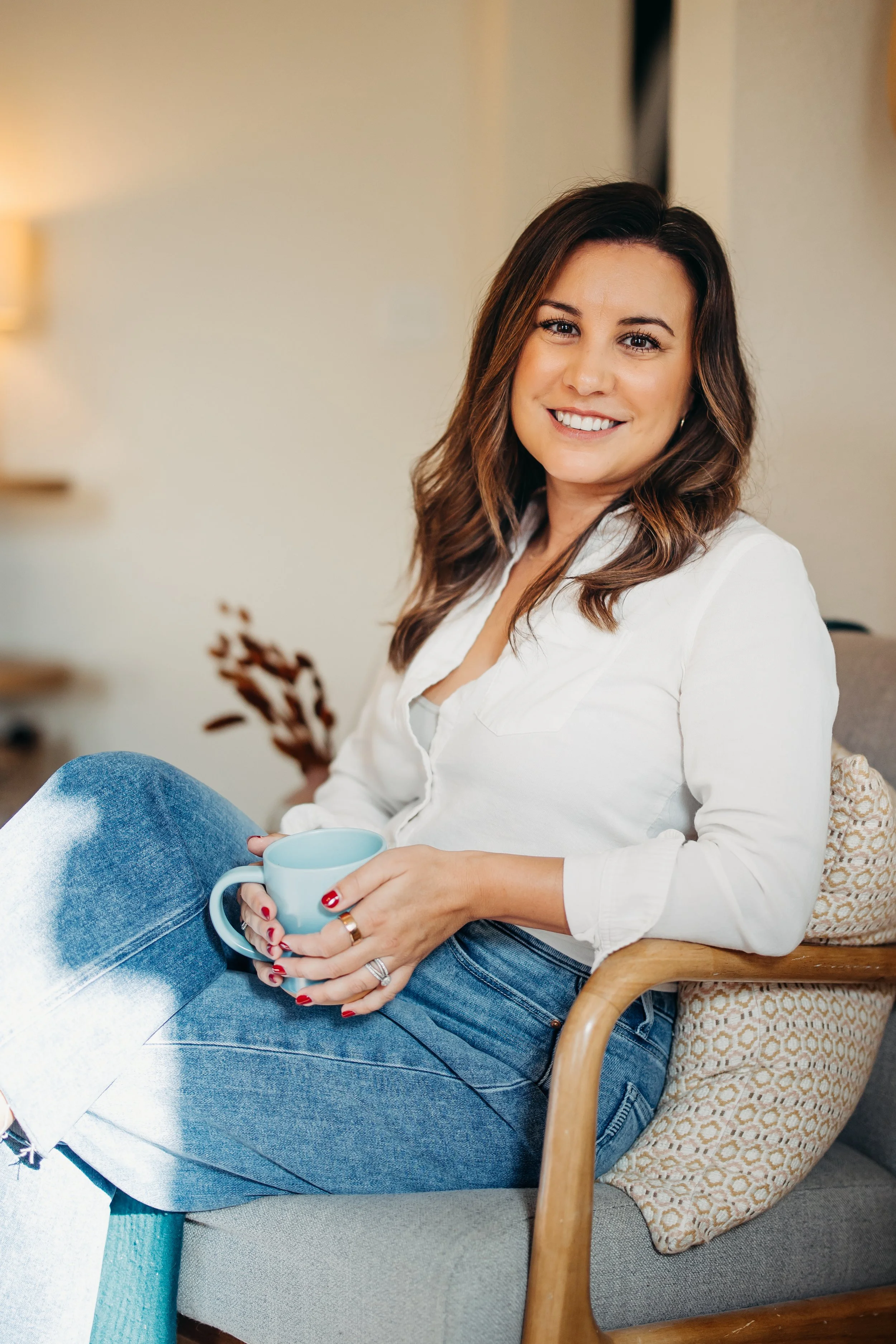 A woman with brown, wavy hair smiling while sitting in a wooden armchair, holding a blue mug, wearing a white blouse and blue jeans.