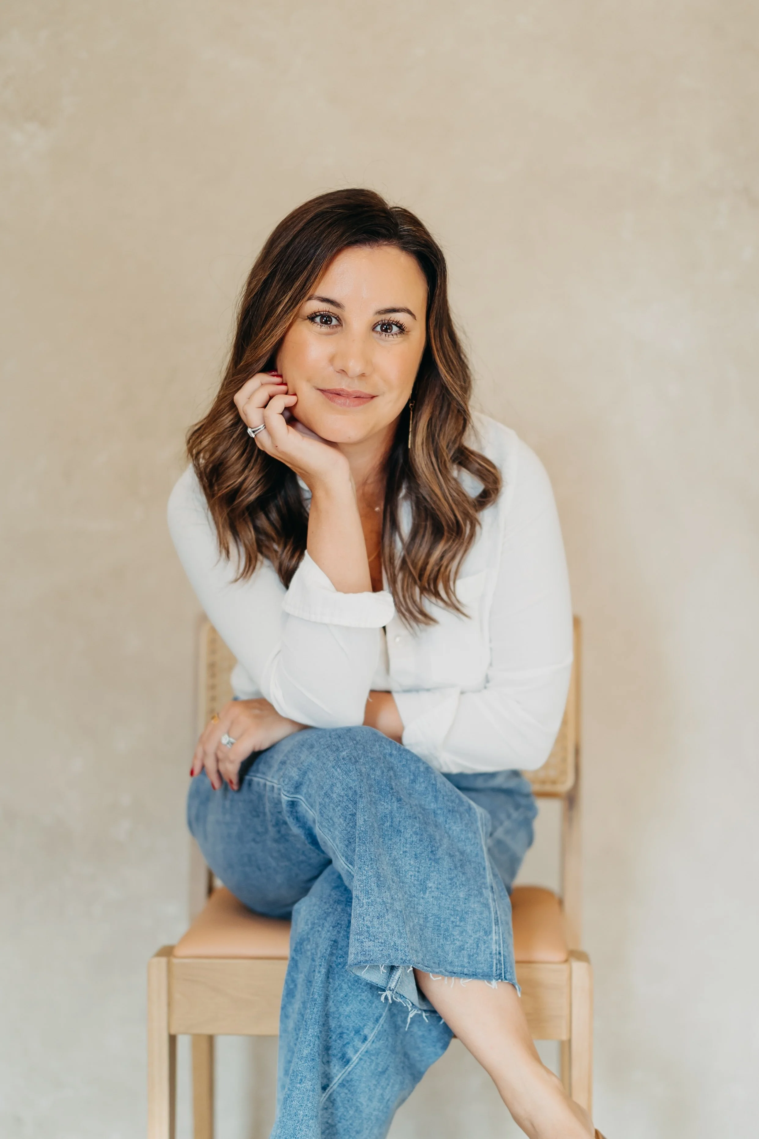 A woman with brown wavy hair wearing a white blouse and blue jeans, sitting on a wooden chair against a beige wall, smiling gently with her chin resting on her hand.