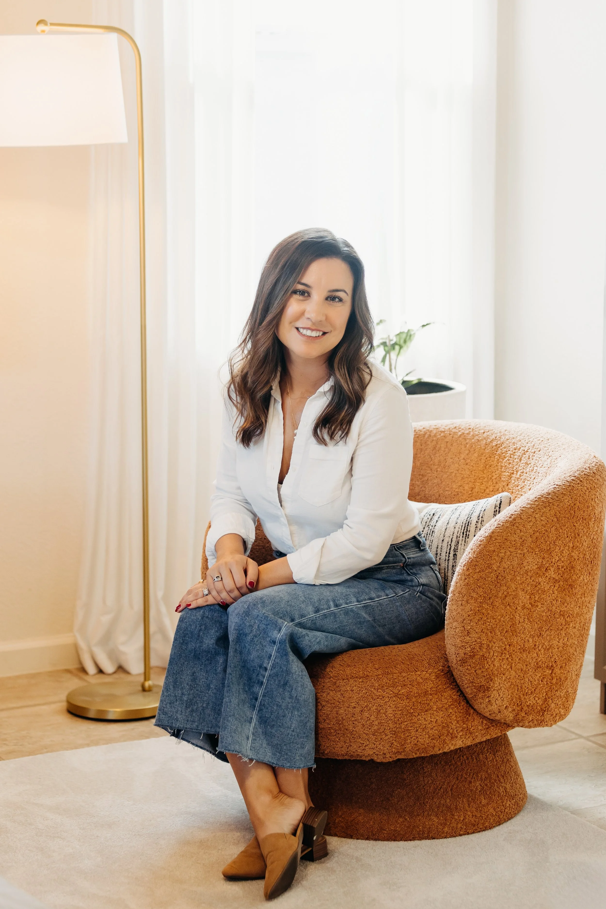 A woman with dark brown hair sitting on a round, orange upholstered chair near a white wall and window, smiling at the camera.