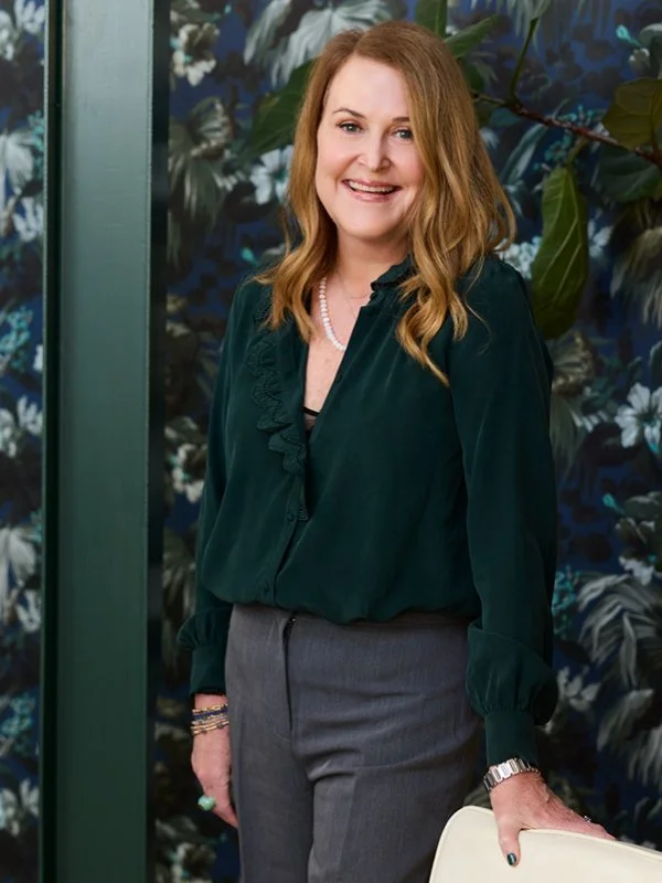A picture of Lisa Maloof, a woman with long red hair, smiling, wearing a dark green blouse, gray pants, and jewelry, standing in front of a dark floral wallpaper background.