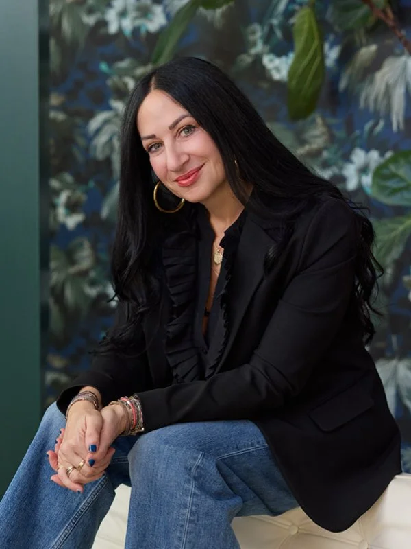 A picture of Wendy Lowden, a woman with long black hair, wearing a black blazer, blue jeans, and gold hoop earrings, sitting on a white cushioned seat, against a background of green leaves and flowers.