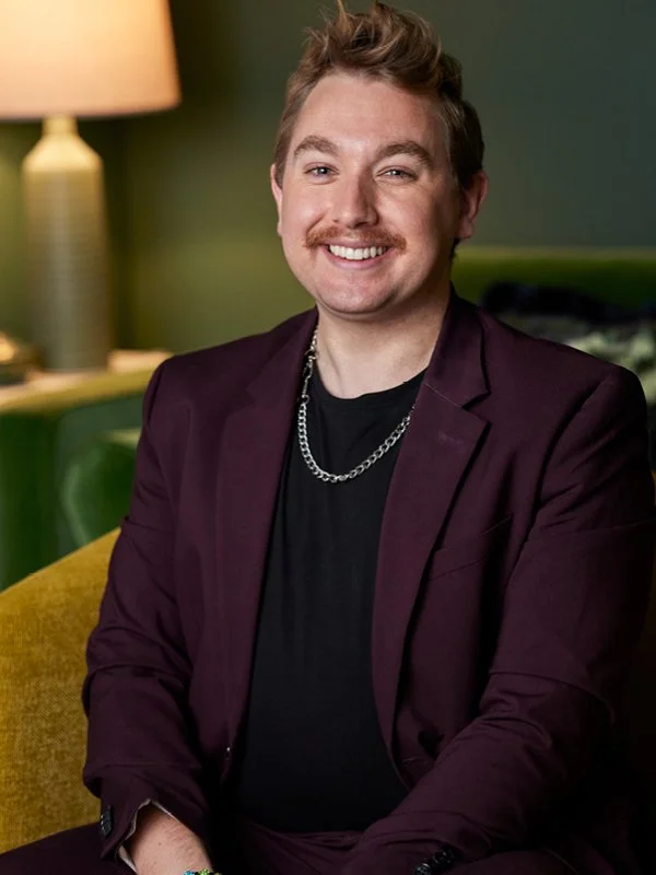 Grayson Hester; Man sitting on a chair, smiling, with a lamp and green wall in the background.
