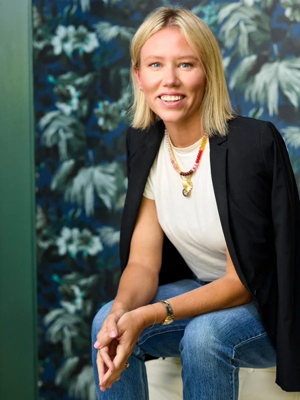 Stefani Demoff; A woman with blonde hair wearing a white t-shirt, a black blazer draped over her shoulders, blue jeans, and accessories including a necklace and a bracelet, sitting in front of a leafy patterned background and smiling at the camera.