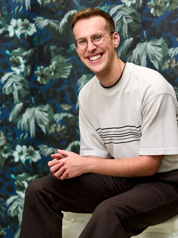 Nick Milavec; A young man with glasses, smiling, sitting on a beige ottoman against a tropical-patterned wall with green leaves and white flowers, wearing a white t-shirt and black pants.
