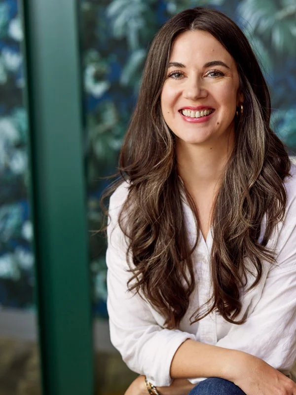 Sierra Serafica; A woman with long brown hair smiling, wearing a white blouse, sitting in front of a wall.