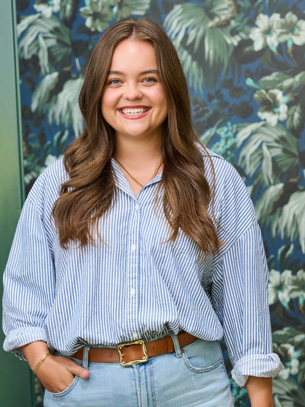 Katie Stone; A young woman with long brown hair and blue eyes smiling, wearing a blue and white striped shirt, blue jeans, and a brown belt, standing in front of a dark leafy floral background.