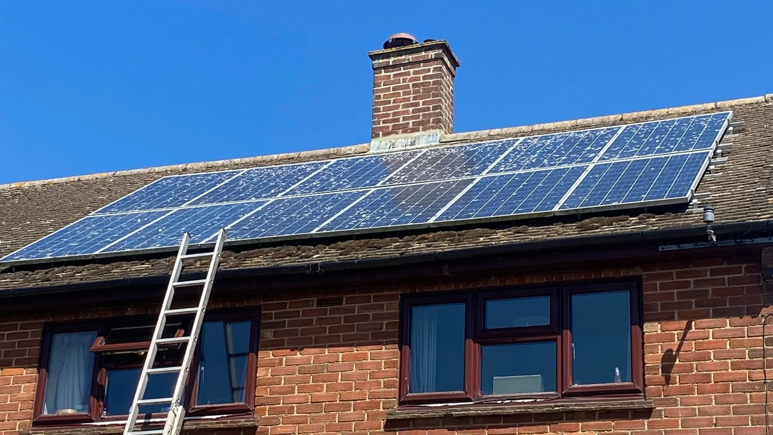 A brick house with solar panels installed on the roof, chimney, blue sky, and a ladder leaning against the house.