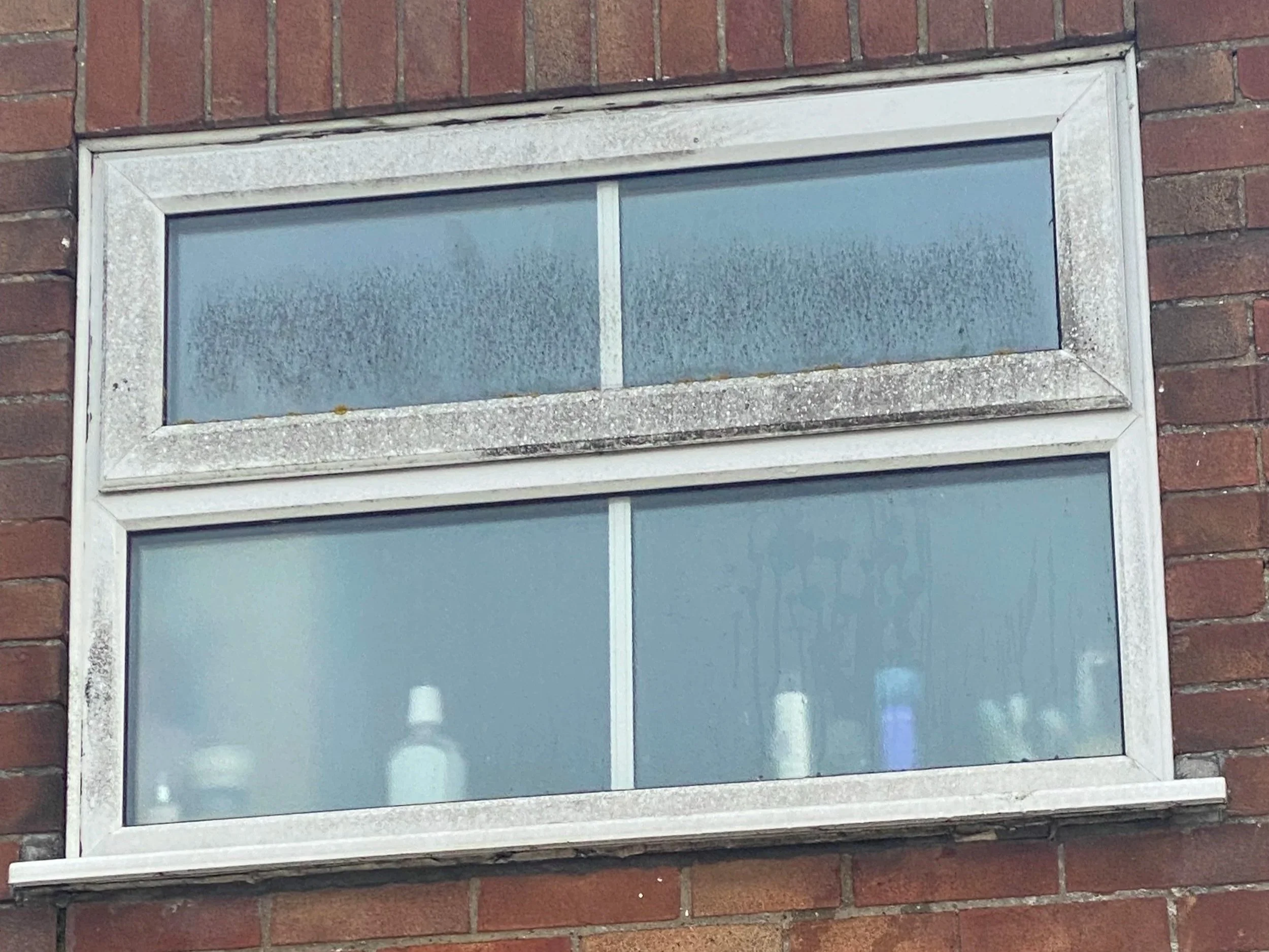 A rectangular basement window with frosted glass panes, set in a brick wall, with a white frame and frosted glass.