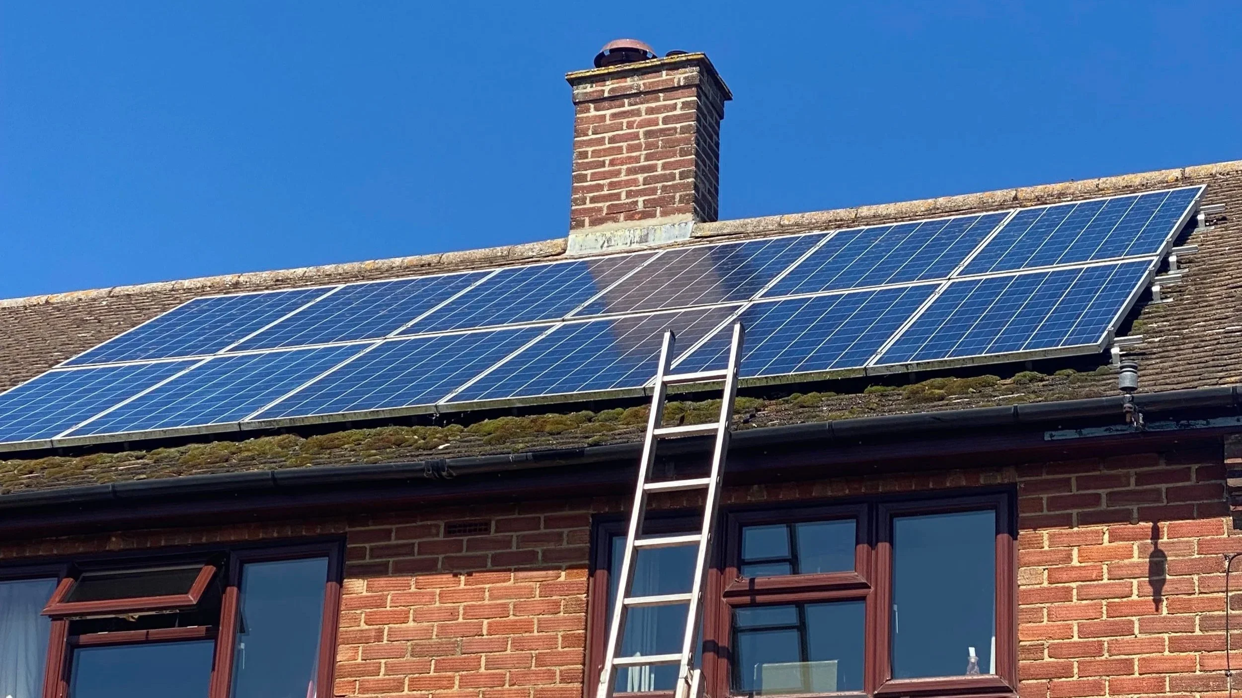 Solar panels installed on a brick house roof with a chimney in the background and a ladder leaning against the house.