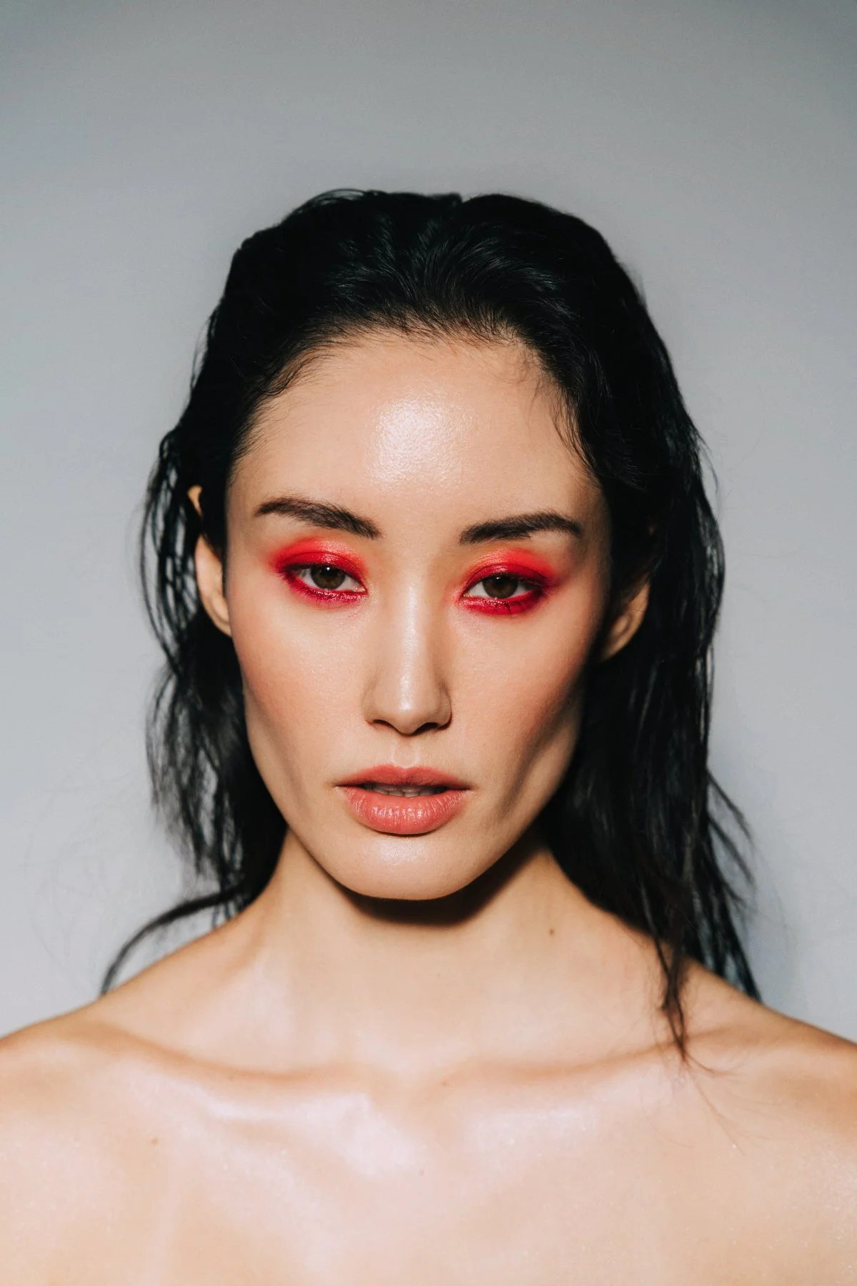 Close-up of a woman with wet black hair, pale skin, and with bright red eyeshadow, looking at the camera against a plain background.