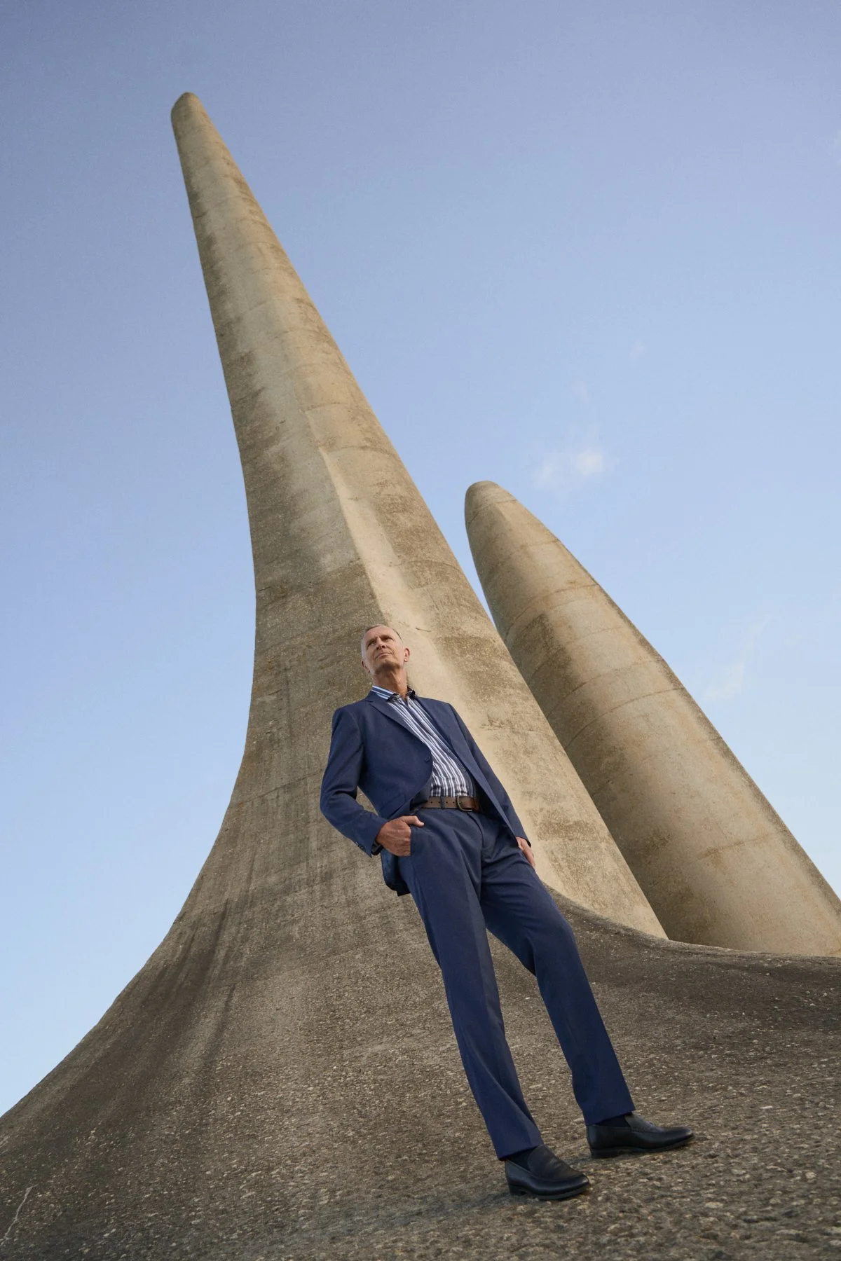 A man in a blue suit standing with hands in pockets in front of large concrete structures that resemble whale fins, with a clear blue sky in the background.
