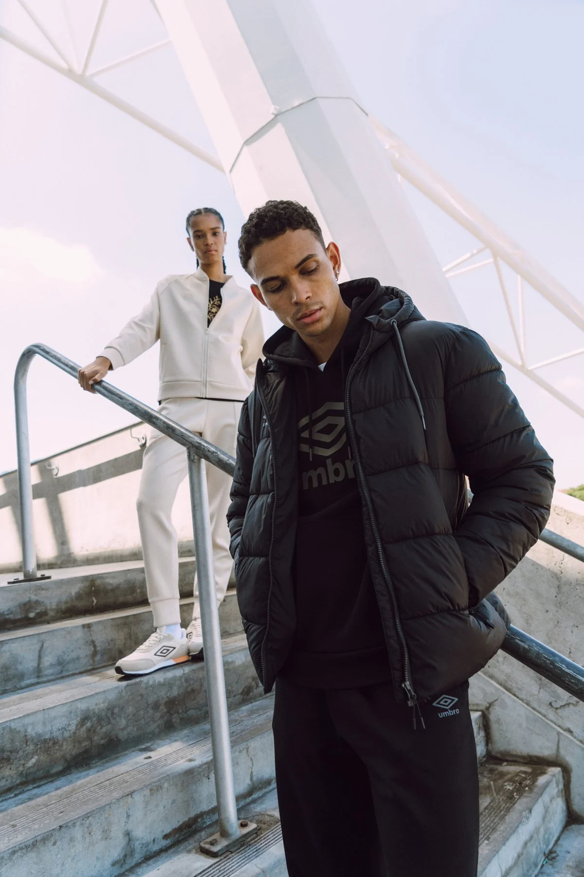 Two young adults, a man and a woman, standing on outdoor concrete stairs near a metal railing, against a clear sky and modern structure. The man is in the foreground, looking down, wearing a black puffer jacket and black hooded sweatshirt. The woman 