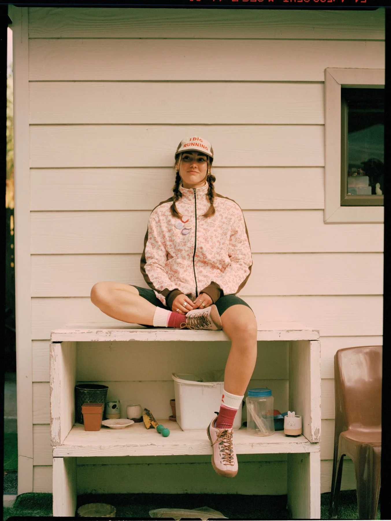 A young woman with braided hair sitting on a white wooden bench against a white wall, wearing a pink patterned jacket, black shorts, pink socks, and sneakers, with a white cap that says 'DIC RUNNING'.