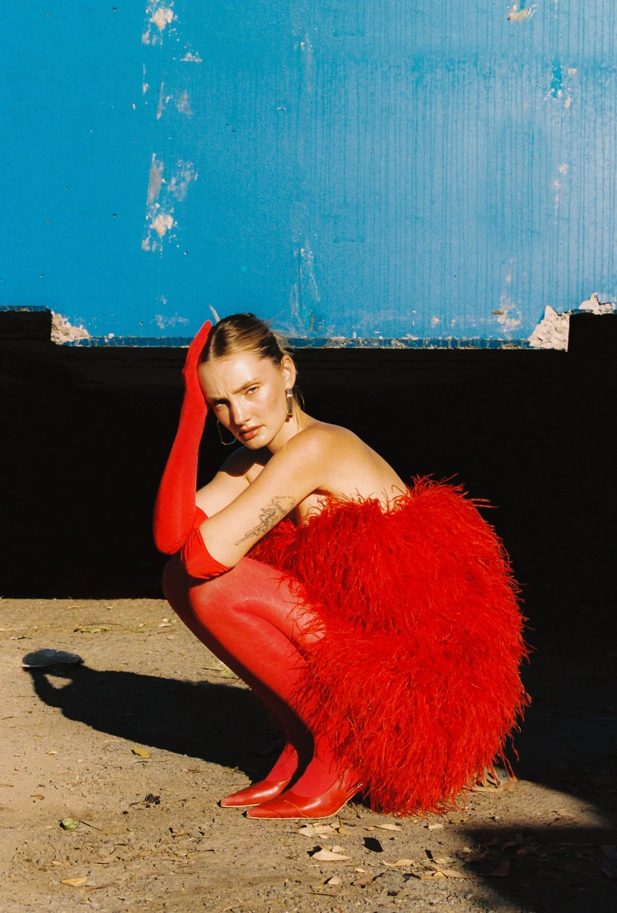 A woman in a dramatic red feathered dress with matching red gloves and high-heeled shoes, crouching on the ground with one hand on her head, in front of a weathered blue wall.