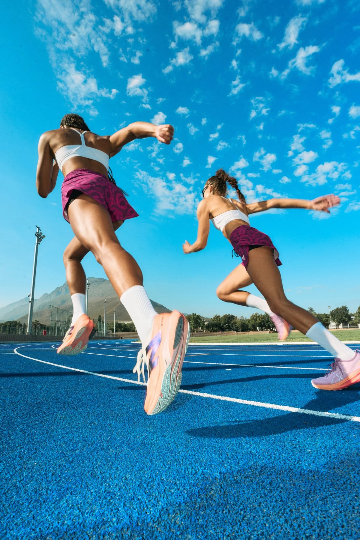 Two female athletes sprinting on a blue running track outdoors with mountains and trees in the background under a partly cloudy sky.