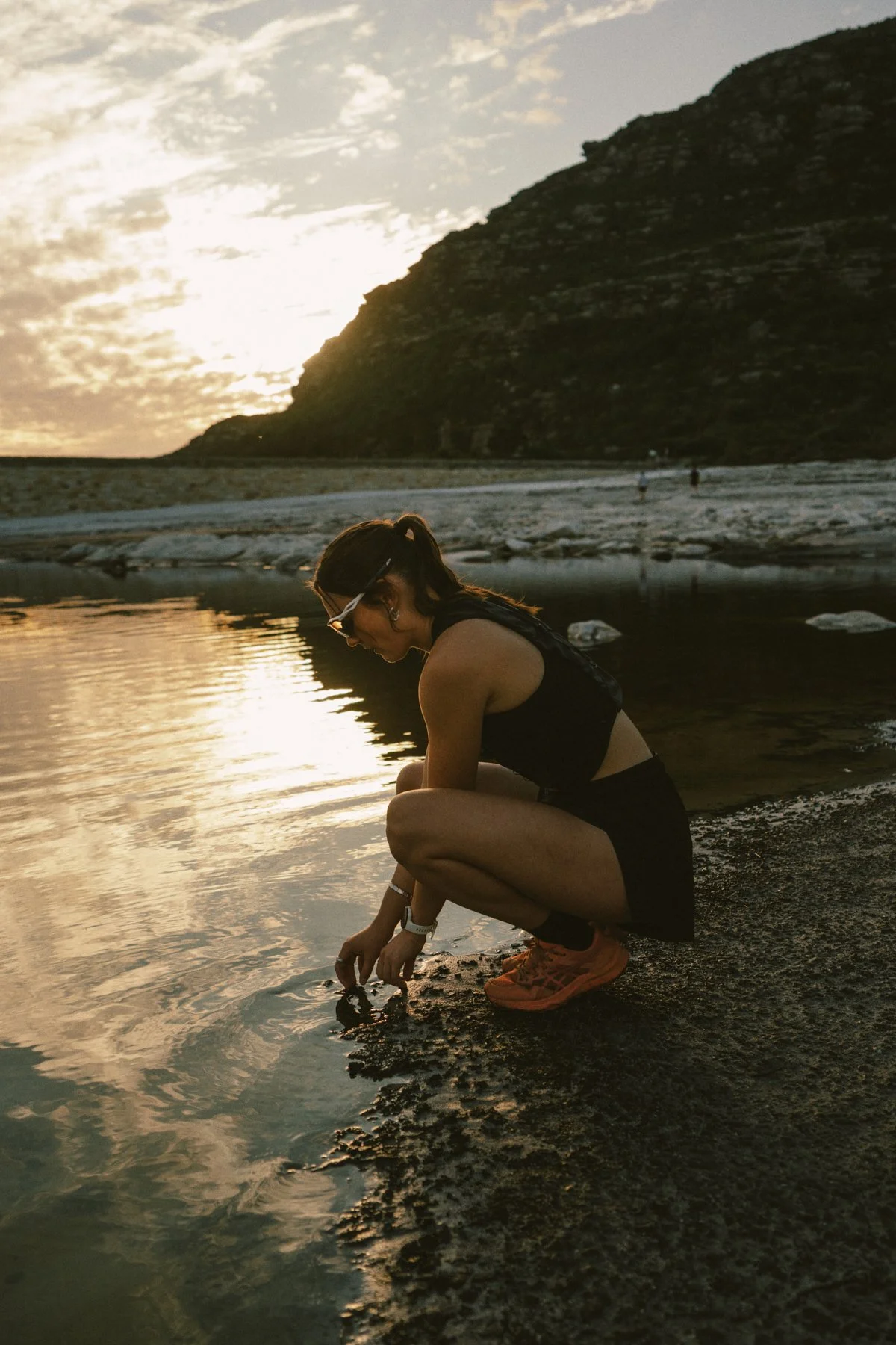 A woman crouching on a rocky shoreline at sunset, touching the water with her hand, wearing sunglasses, a black sleeveless top, a black skirt, and orange sneakers, with a mountain in the background.