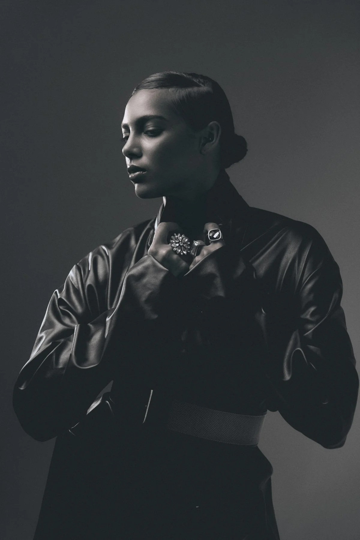 Black and white portrait of a woman with her eyes closed, wearing a leather jacket and multiple rings, with her hands near her neck, head tilted slightly downward, and her hair styled in a sleek, low bun.