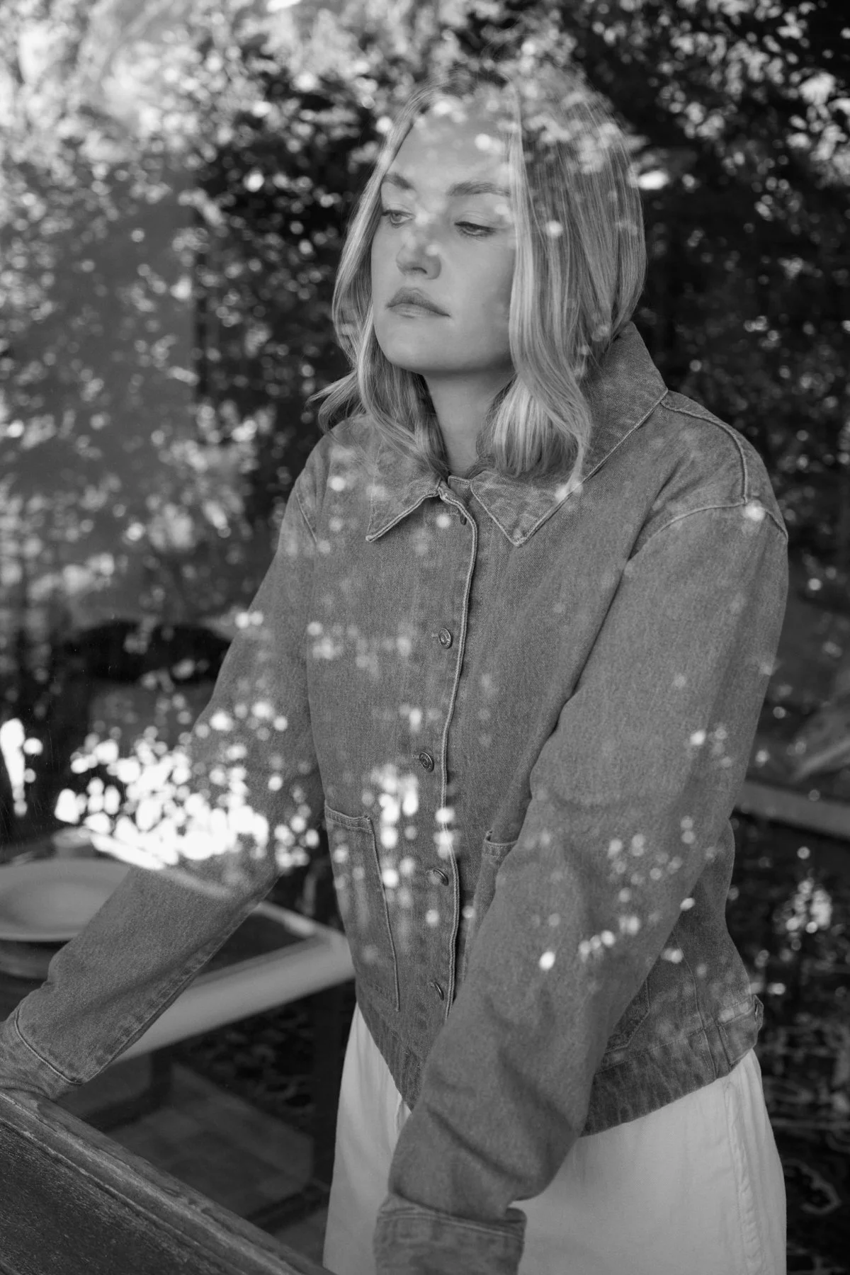 A young woman with blonde hair and light skin standing outdoors, looking down, wearing a denim jacket, with trees and reflections in the background.