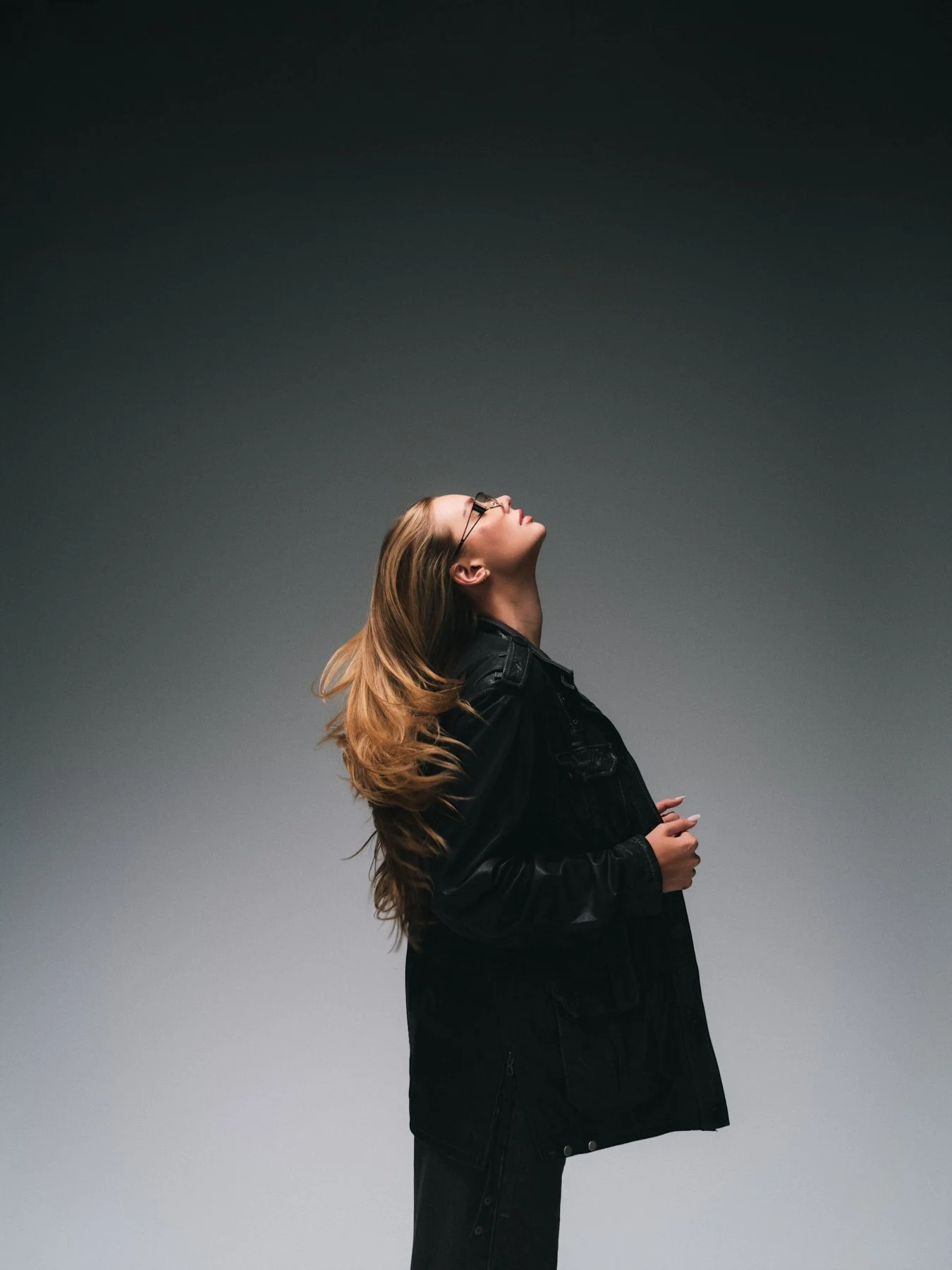A woman with long, wavy, blonde hair wearing glasses and a black leather jacket poses against a gradient dark background, looking upward with her eyes closed.